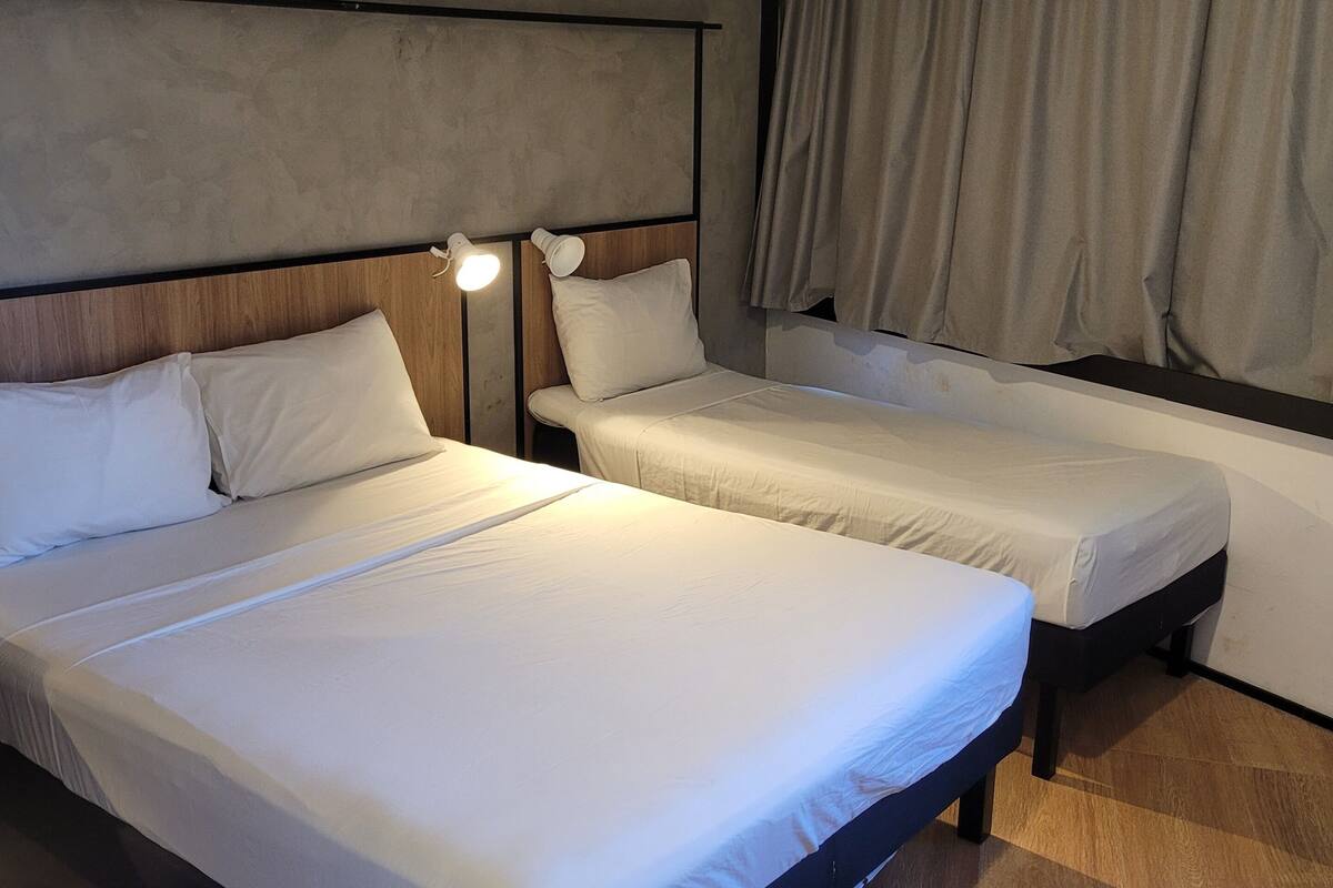 A hotel room with two beds, a headboard, a nightstand with a lamp, and a window with curtains.