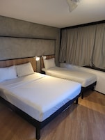 Habitación de hotel con dos camas, cabecera, mesita de noche con lámpara y ventana con cortinas.