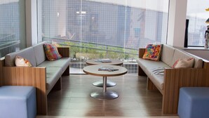 A modern lounge area with two sofas, a round table, and a view of the city through large windows. - Ibis Maceio Pajucara (Maceió)