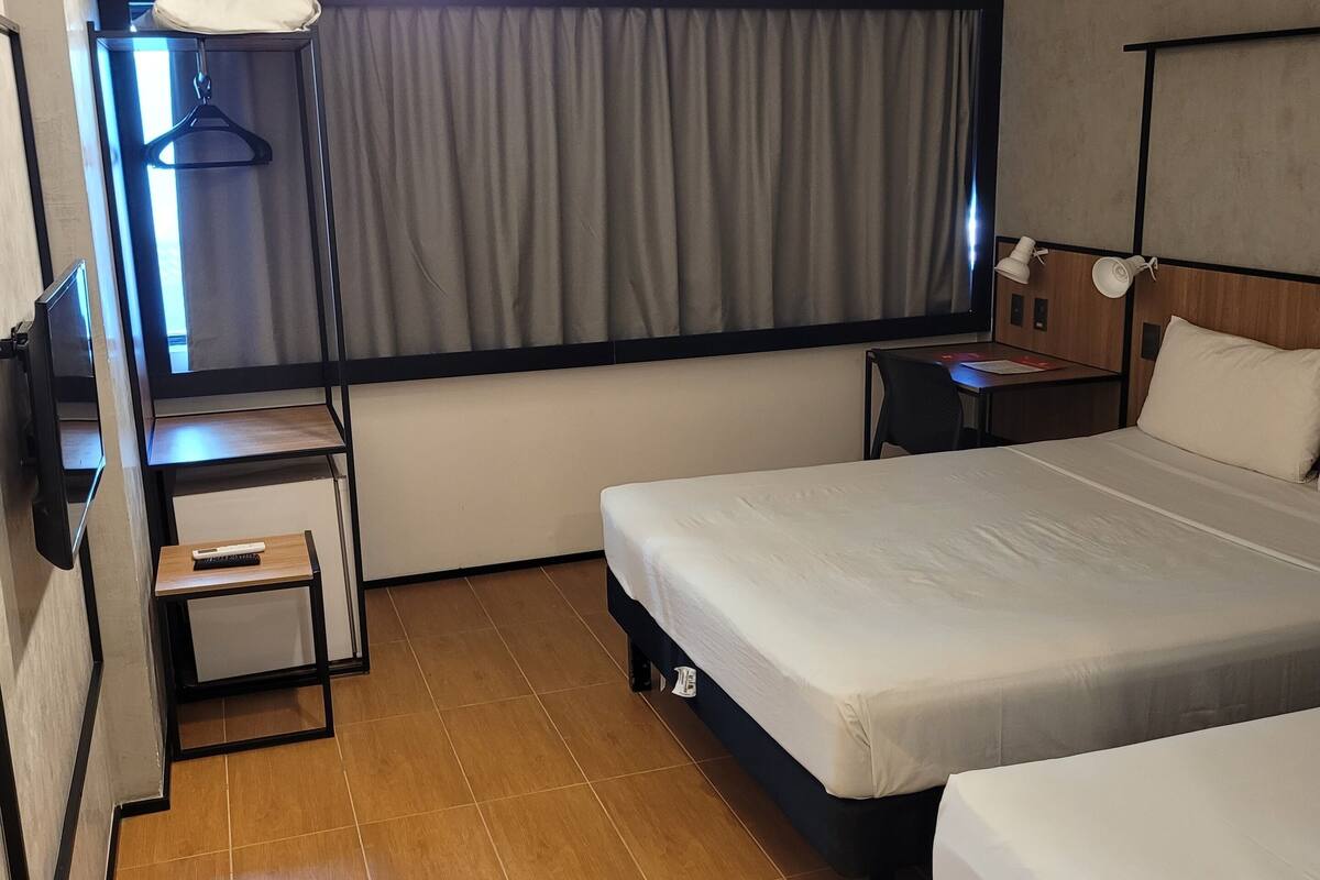 A hotel room with two beds, a desk, a chair, a TV, a lamp, and a window with curtains.