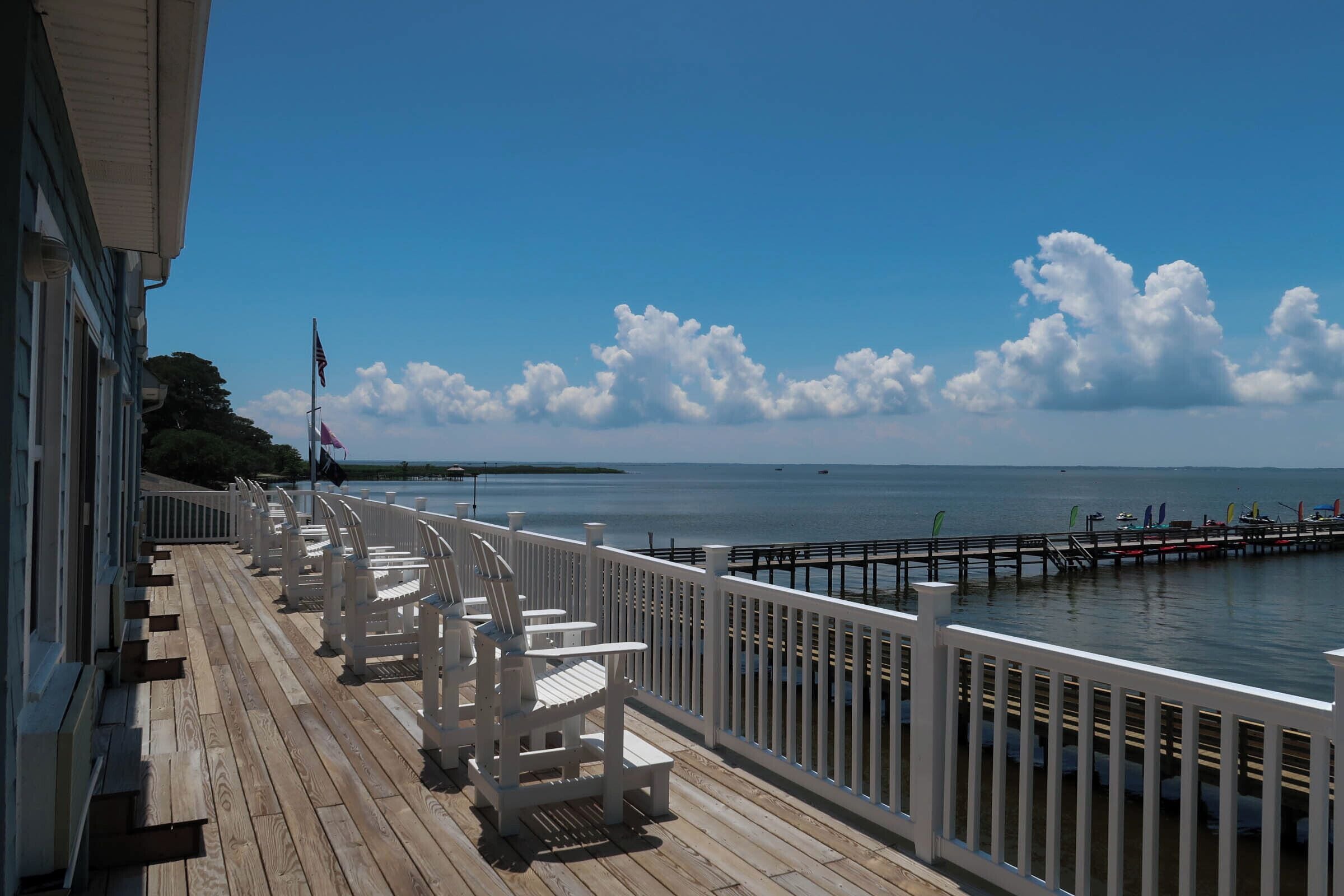 A wooden deck with white chairs and a railing overlooking a body of water.