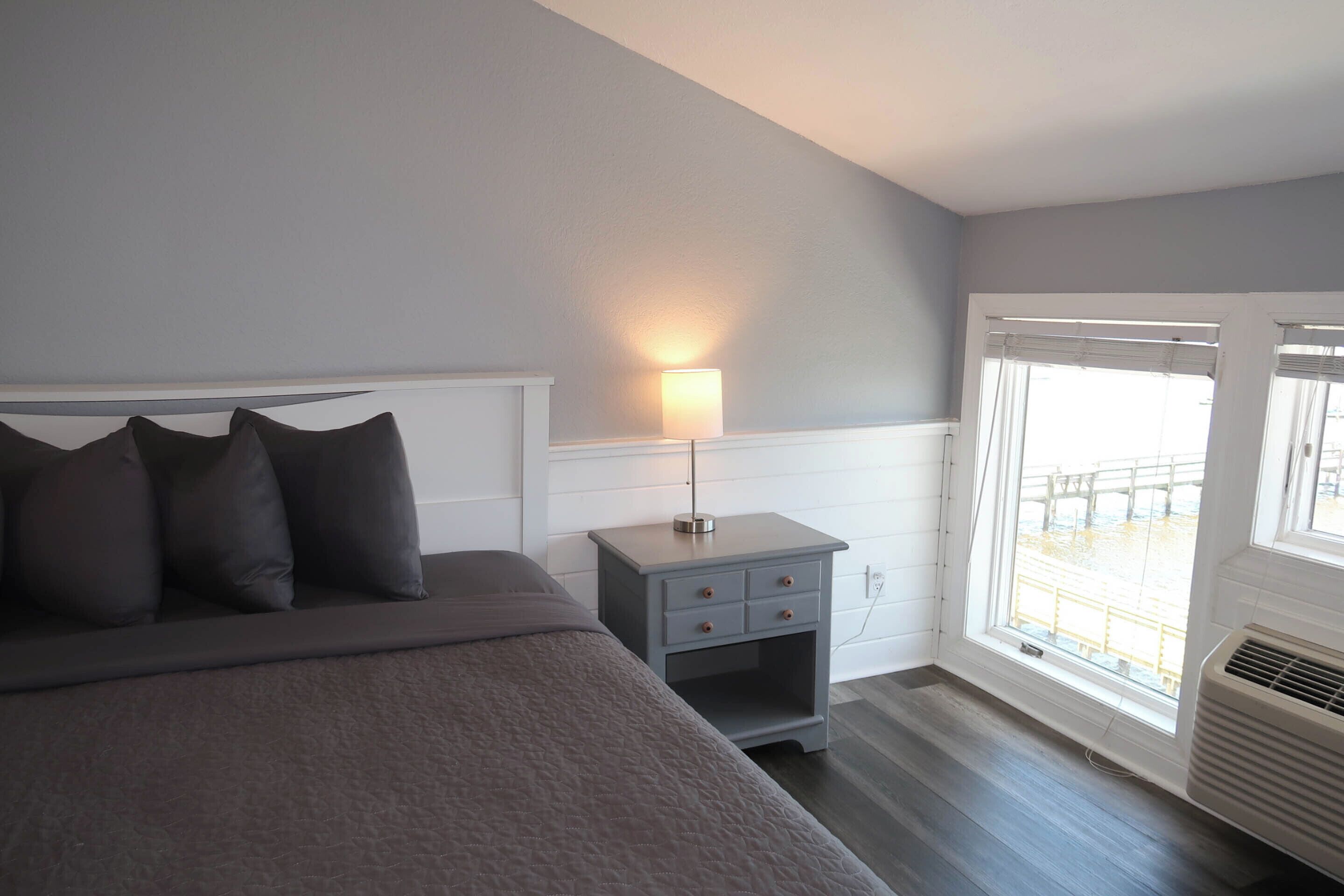 A bedroom with a bed, bedside table, lamp, and a window with a view.