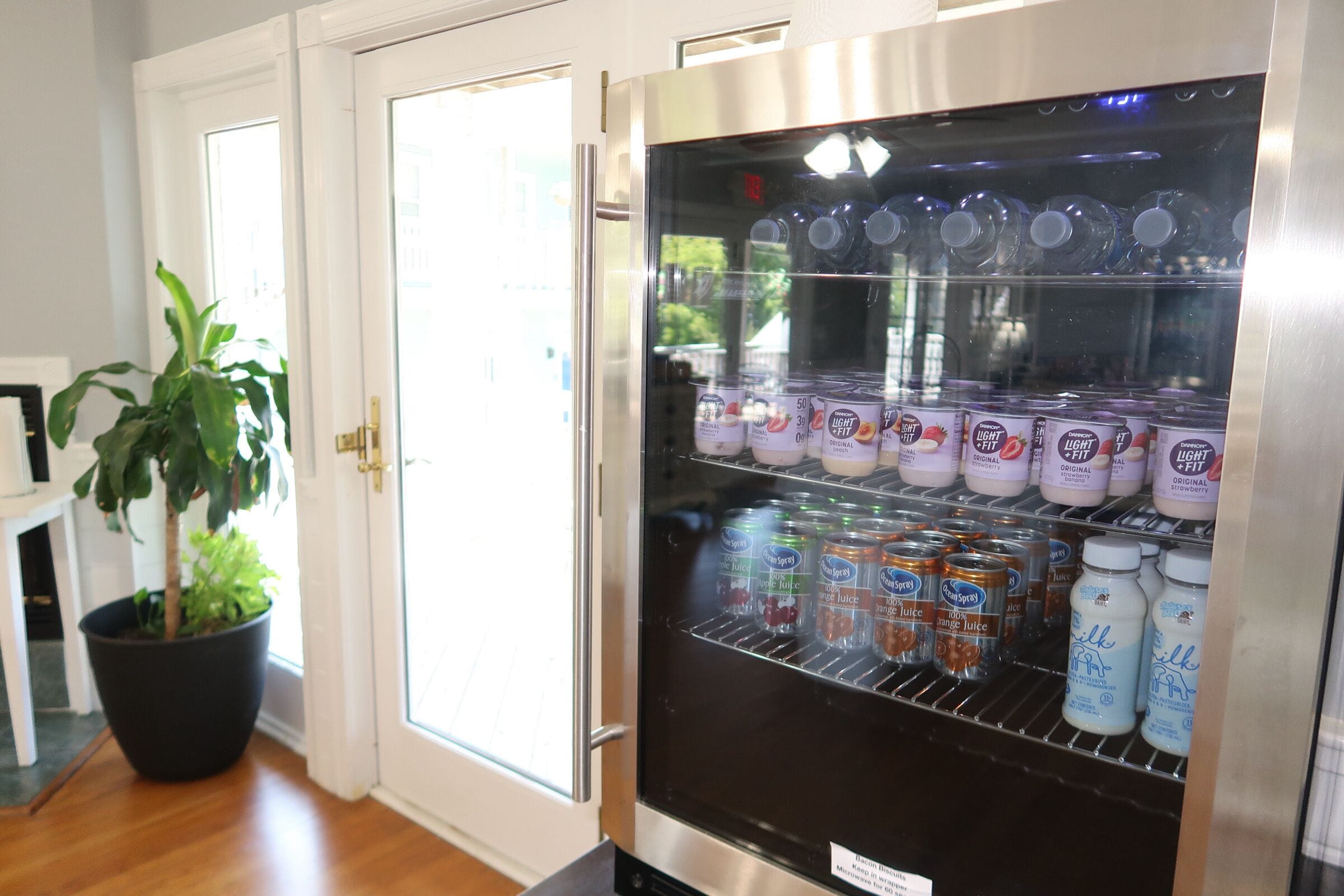 A refrigerator stocked with various beverages, including milk and juice, in a room with wooden flooring and a potted plant.