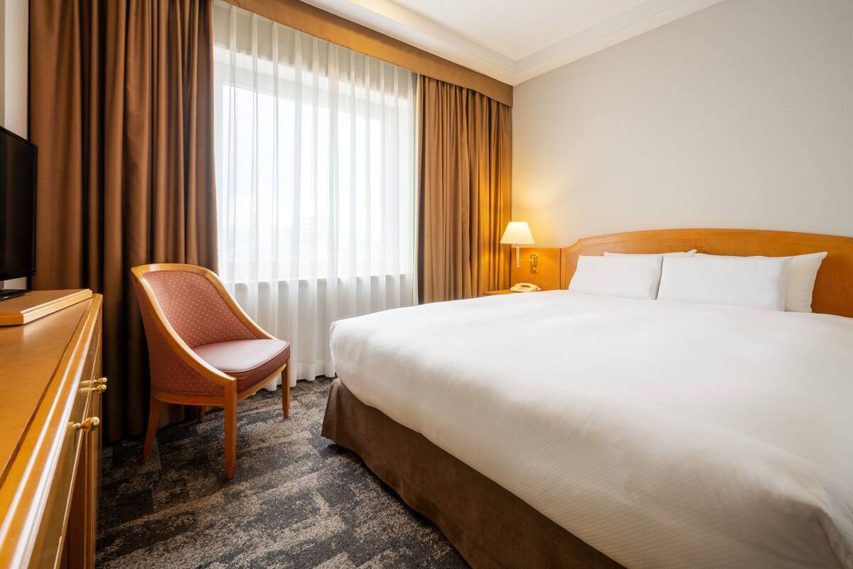 A hotel room with a bed, a chair, a desk with a television, a lamp, and a window with curtains.