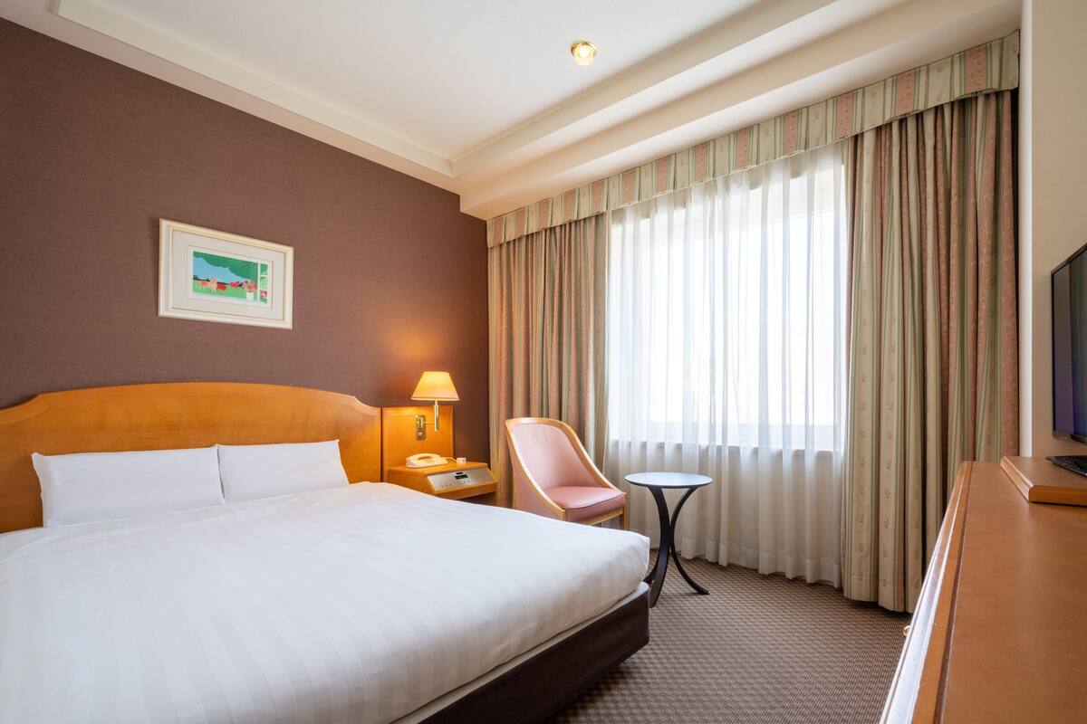 A hotel room with a large bed, a desk, a chair, a small table, a lamp, and a television.