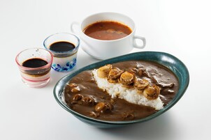 Un tazón de curry con arroz y cebollas fritas, acompañado por dos vasos pequeños de salsa y una taza de té.