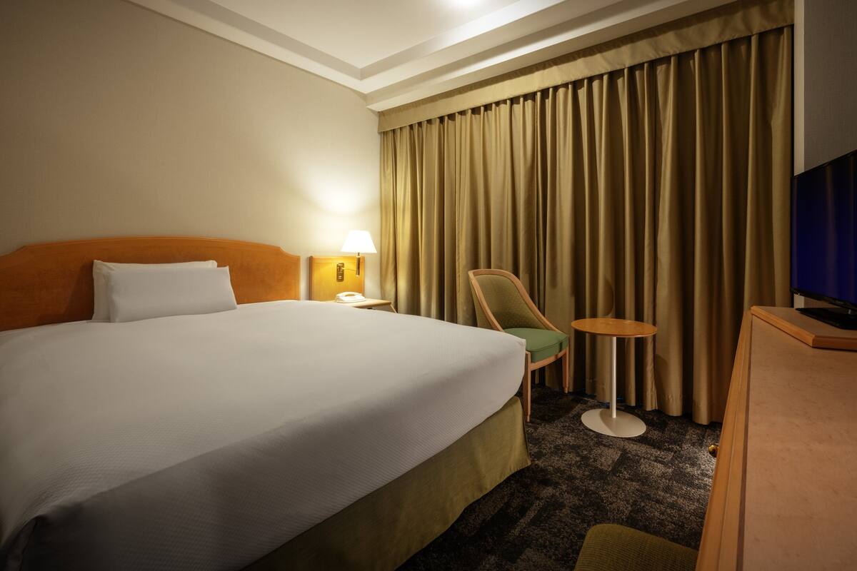A hotel room with a large bed, a desk with a lamp, a chair, and a television.