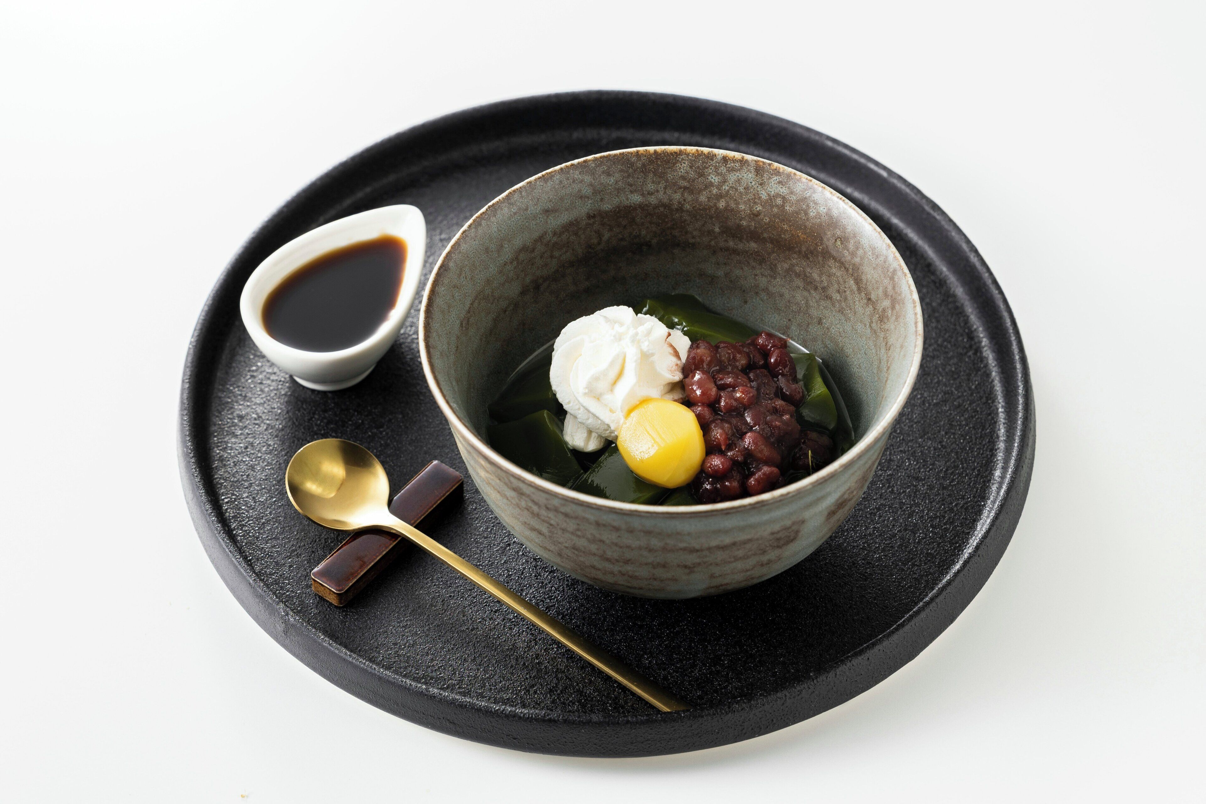 A bowl of dessert with a spoon and a small cup of sauce on a black plate.