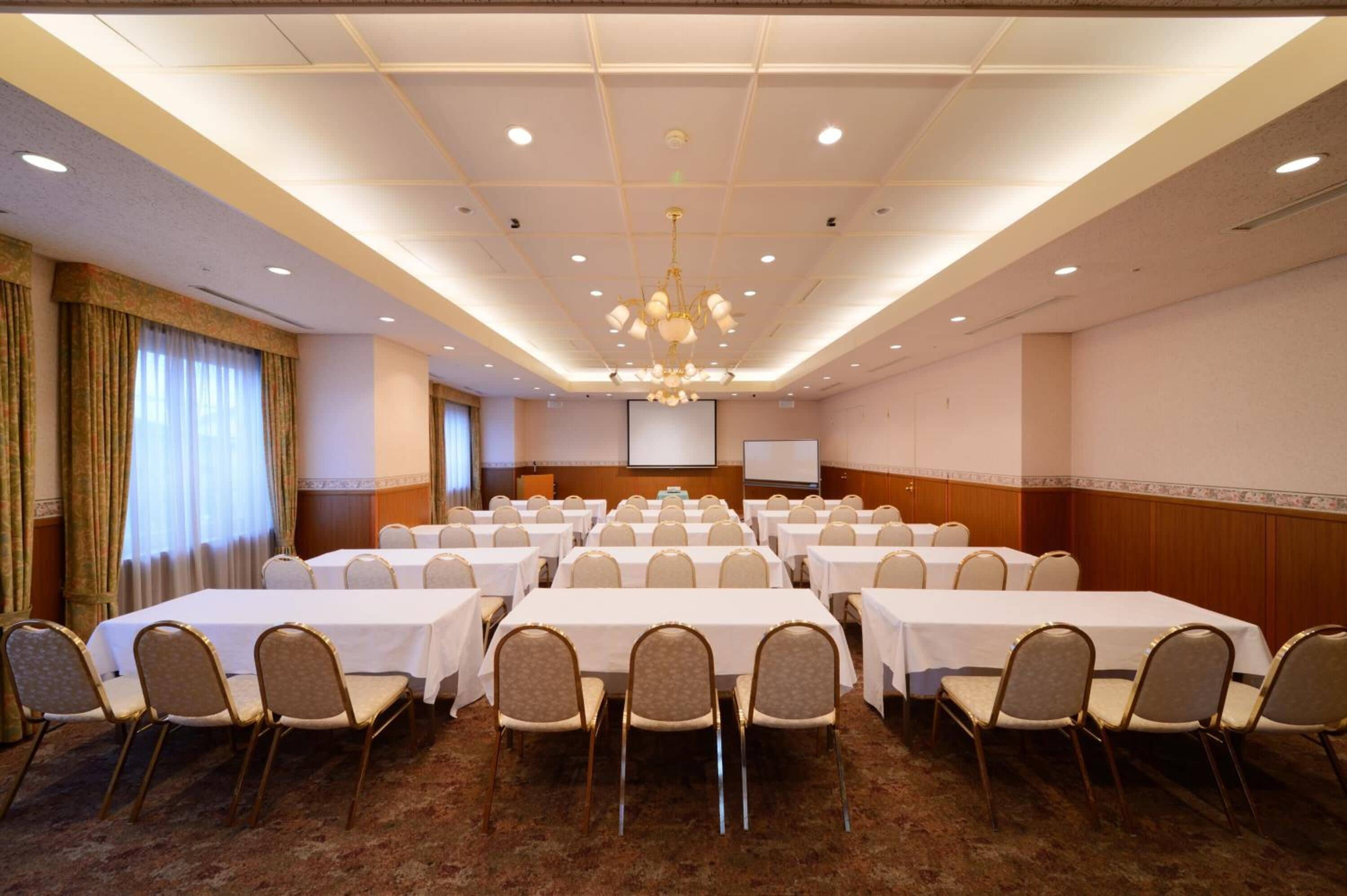 Un salón de conferencias con varias mesas y sillas, una pantalla de proyector y una lámpara de araña.