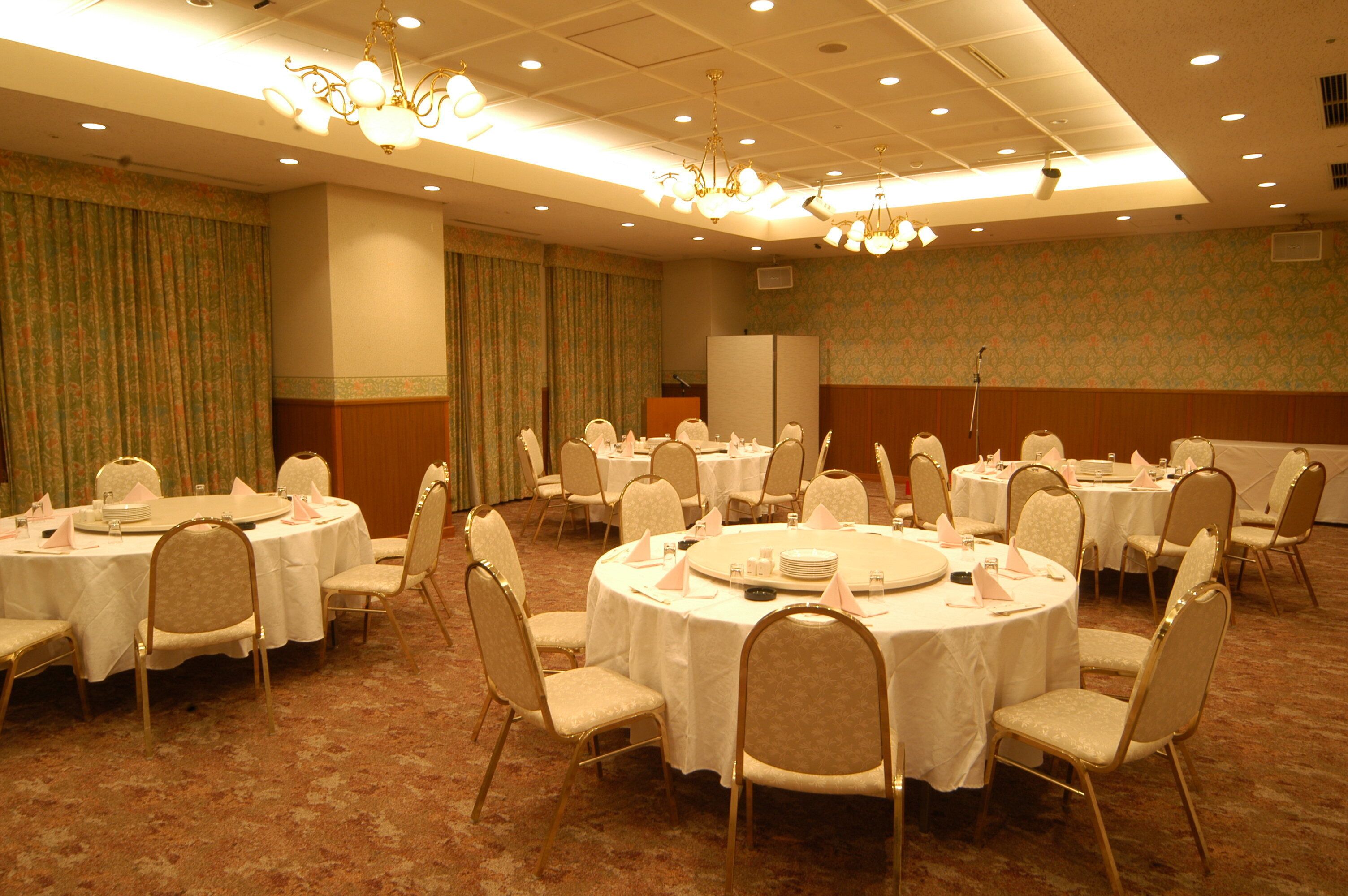 Une salle de conférence dotée de plusieurs tables rondes, chacune recouverte d’une nappe blanche et agrémentée de chaises et de divers éléments de service.