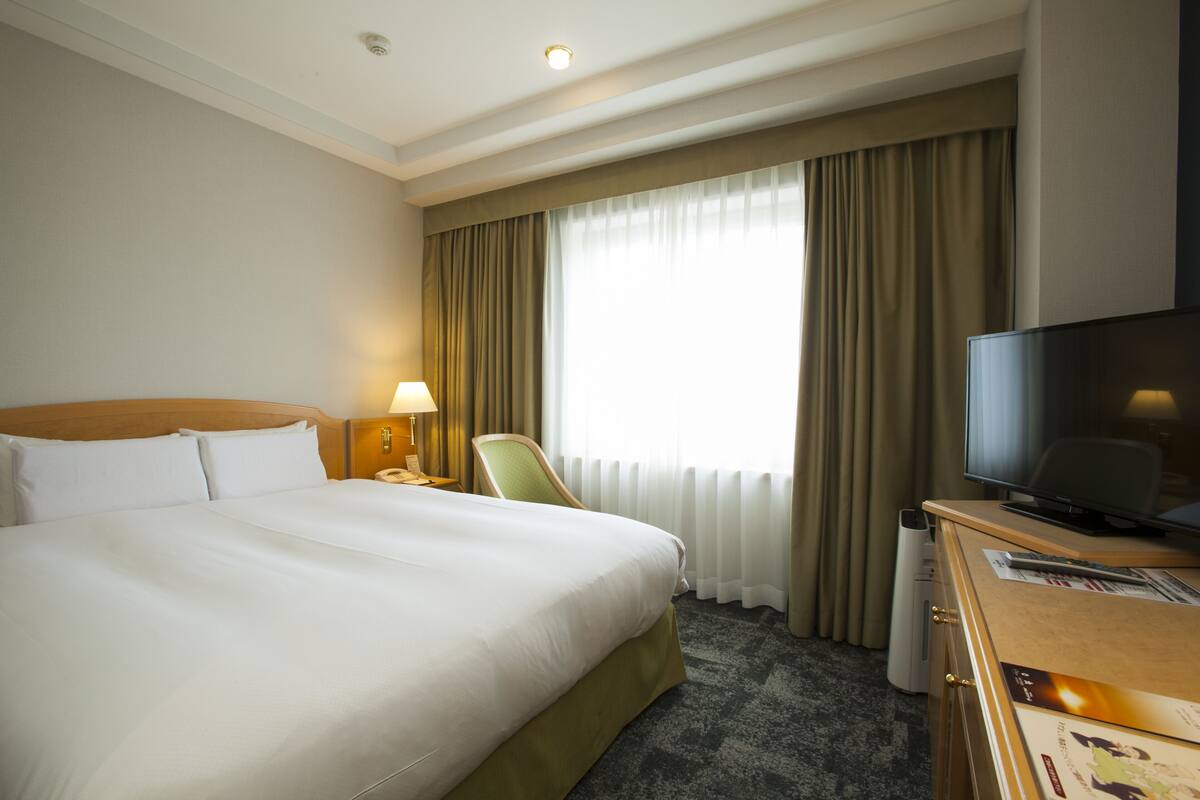 A hotel room with a large bed, a television, a desk with a computer, and a window with curtains.