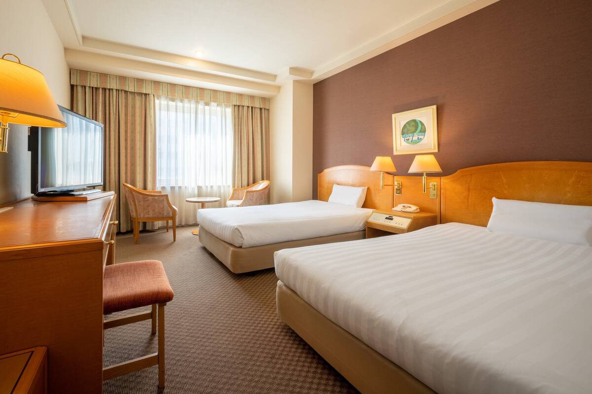 A hotel room with two beds, a desk with a television, a chair, and a small table.