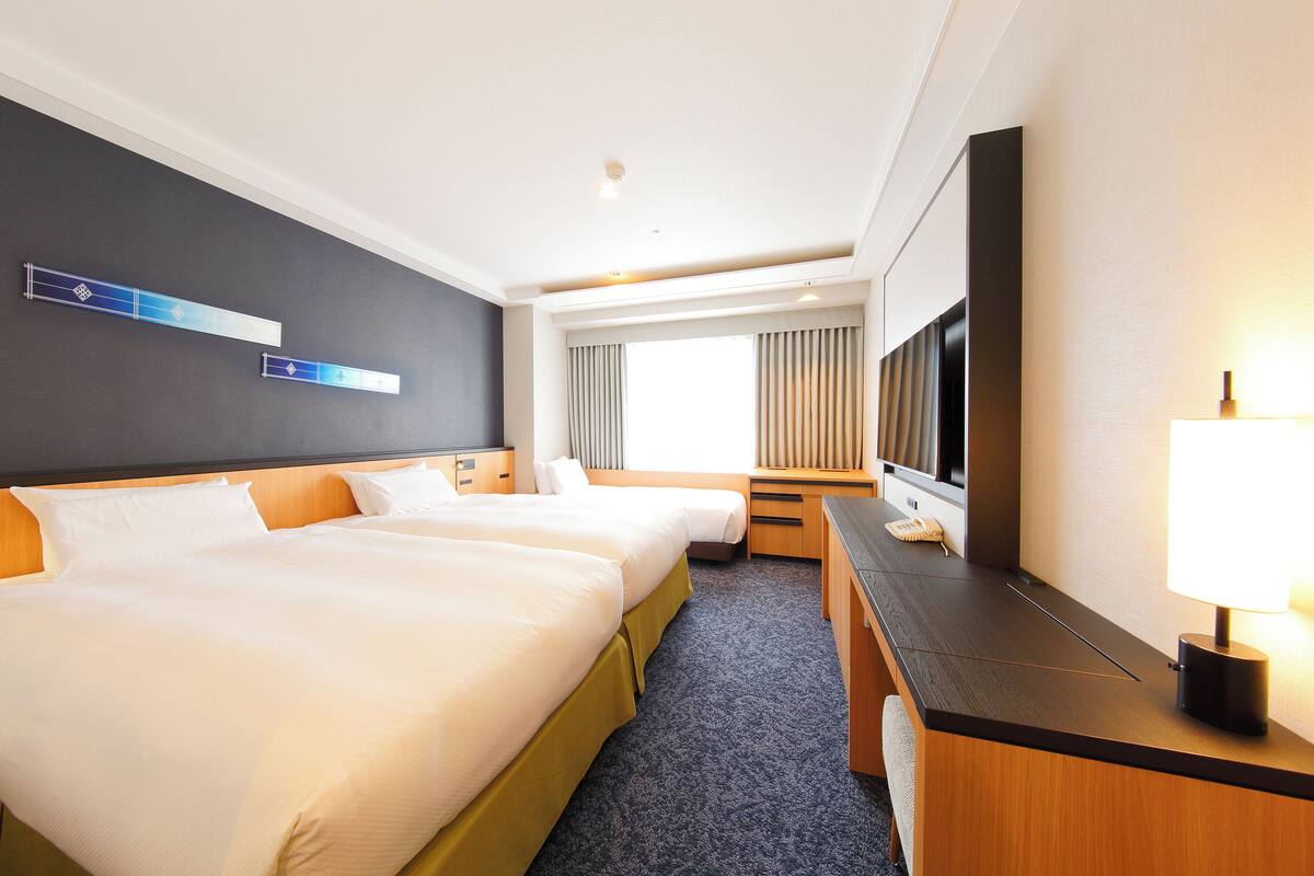 A hotel room with two beds, a desk, a TV, and a window with curtains.
