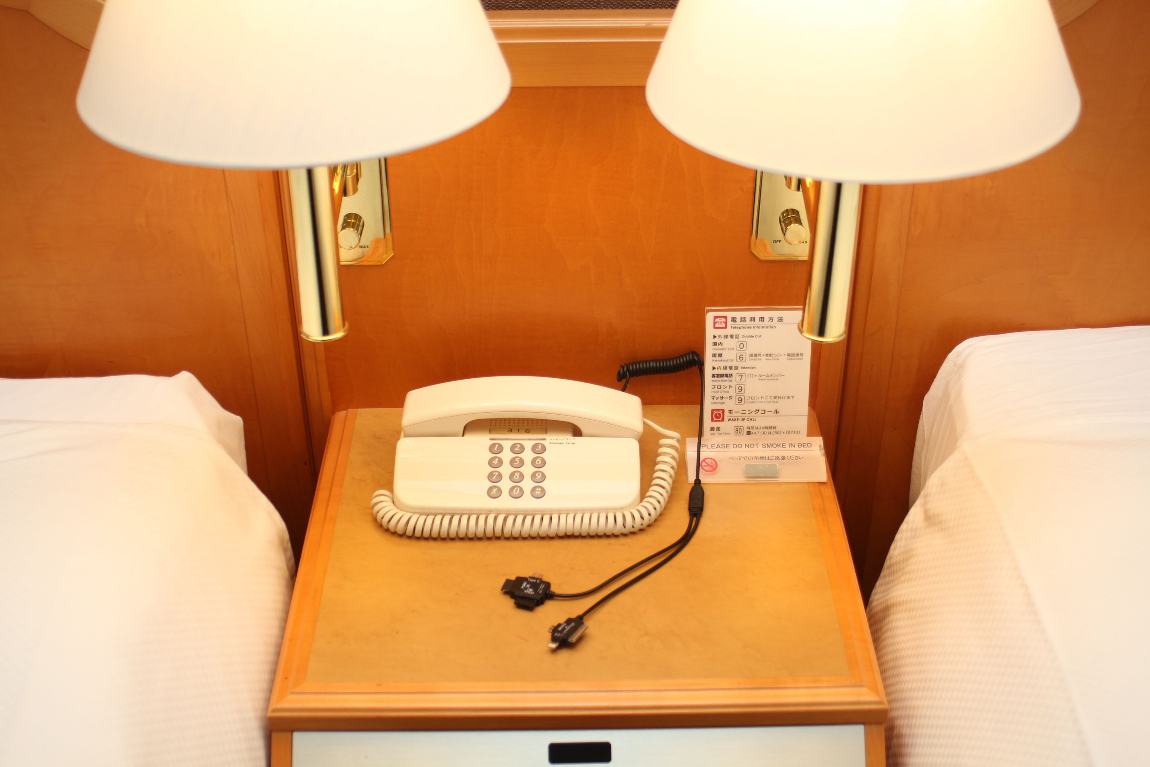 A hotel room with a wooden nightstand, a white telephone, and two lamps.