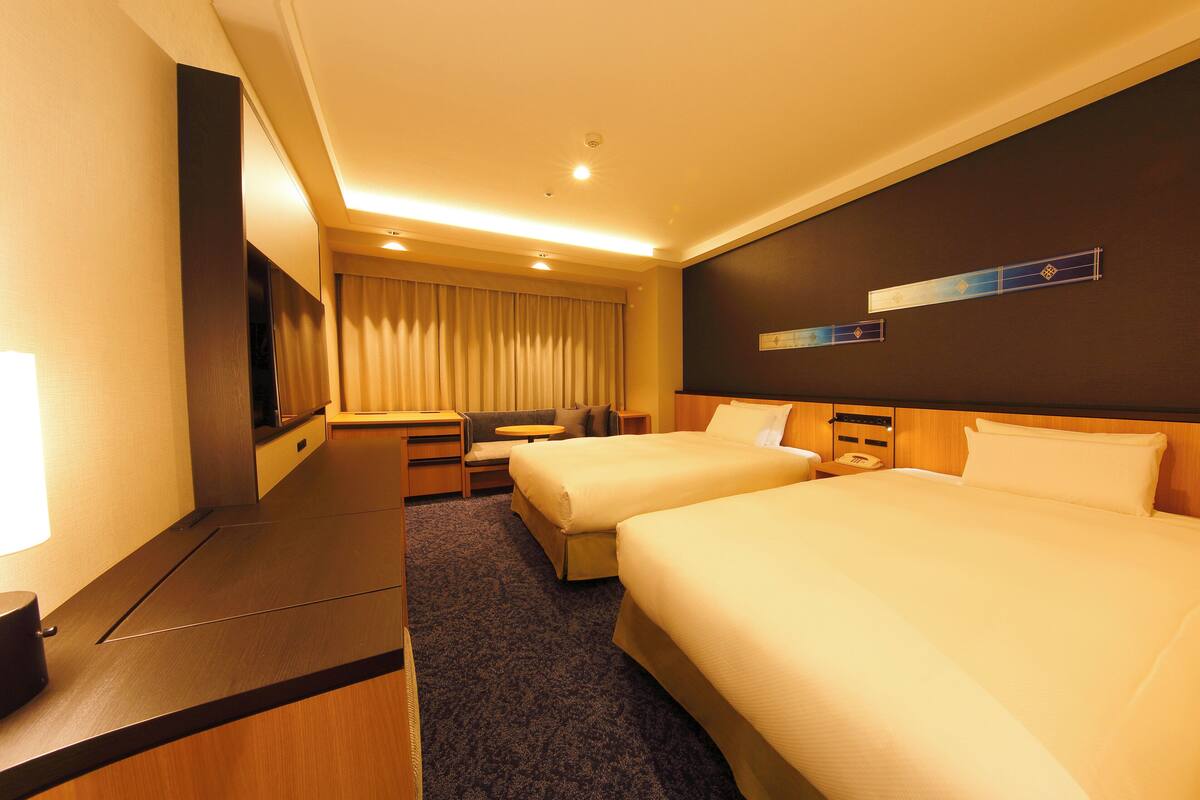 A hotel room with two beds, a desk, a chair, and a TV.