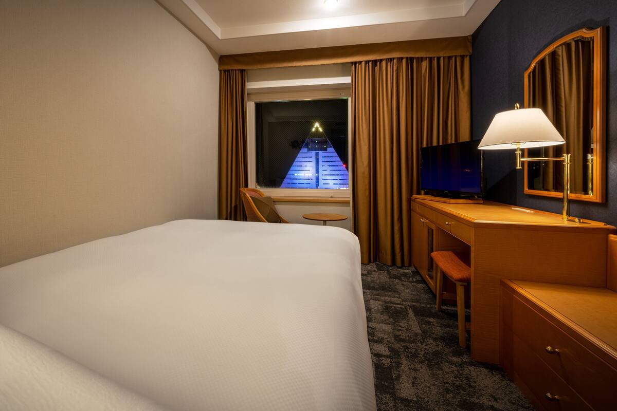 A hotel room with a bed, a desk with a lamp, a television, and a view of a building through the window.