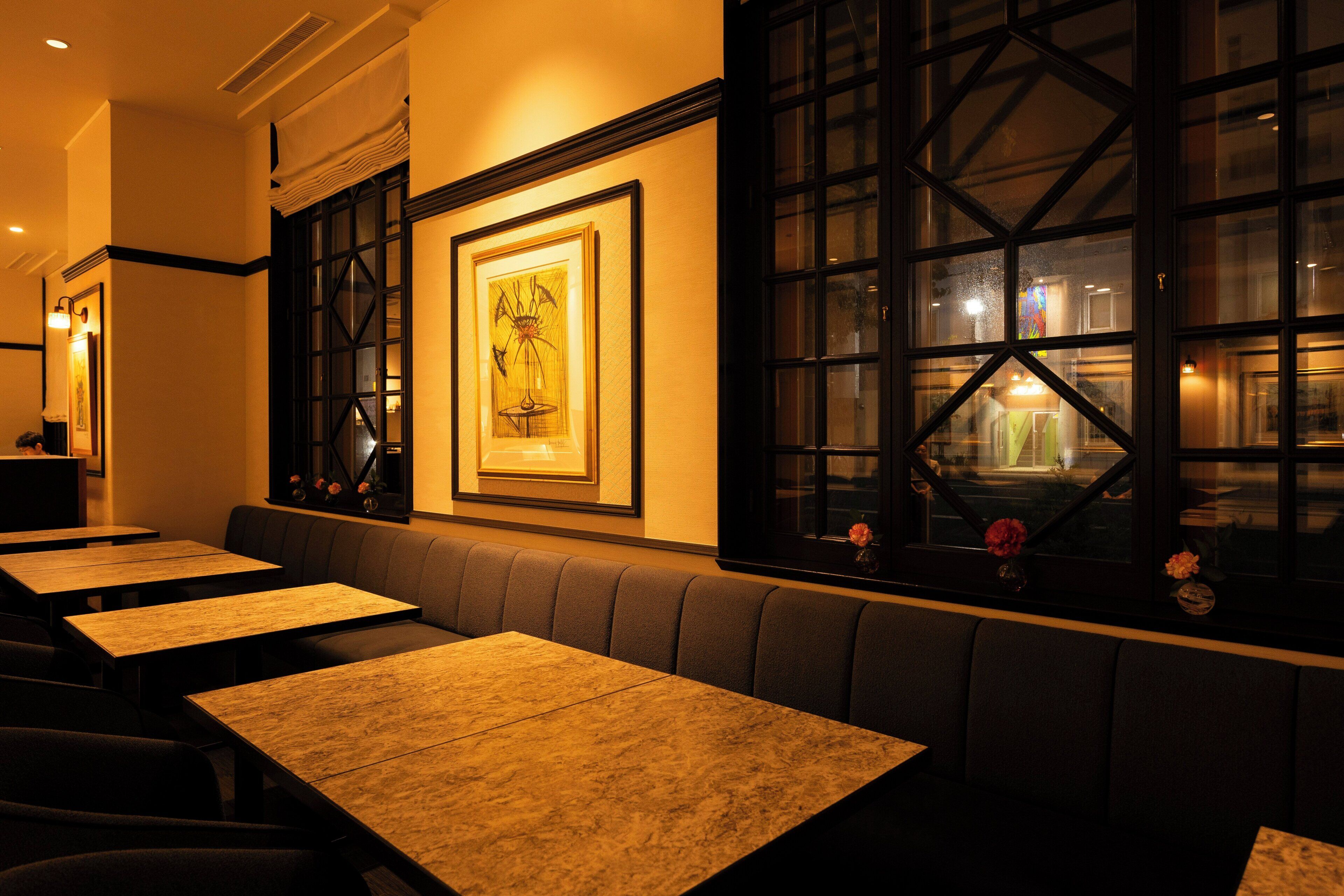 A restaurant interior with wooden tables and benches, framed artwork on the wall, and large windows.