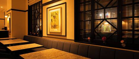 A restaurant interior with wooden tables and benches, framed artwork on the wall, and large windows.