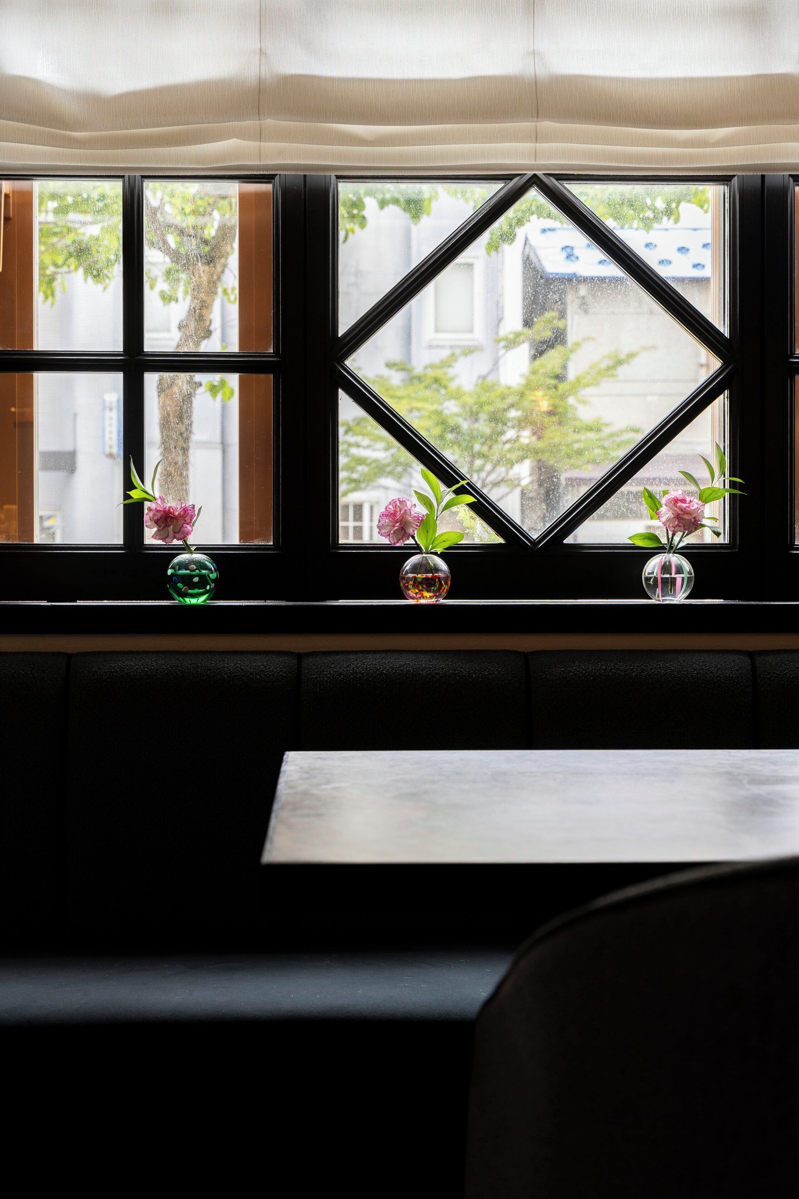 A window with a geometric pattern, three vases with flowers, and a view of a building and trees.