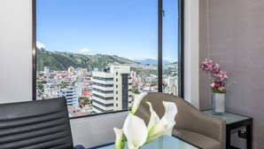 Habitación de hotel con un ventanal grande que ofrece vistas a la ciudad, una mesa de vidrio, una silla y un jarrón con flores.