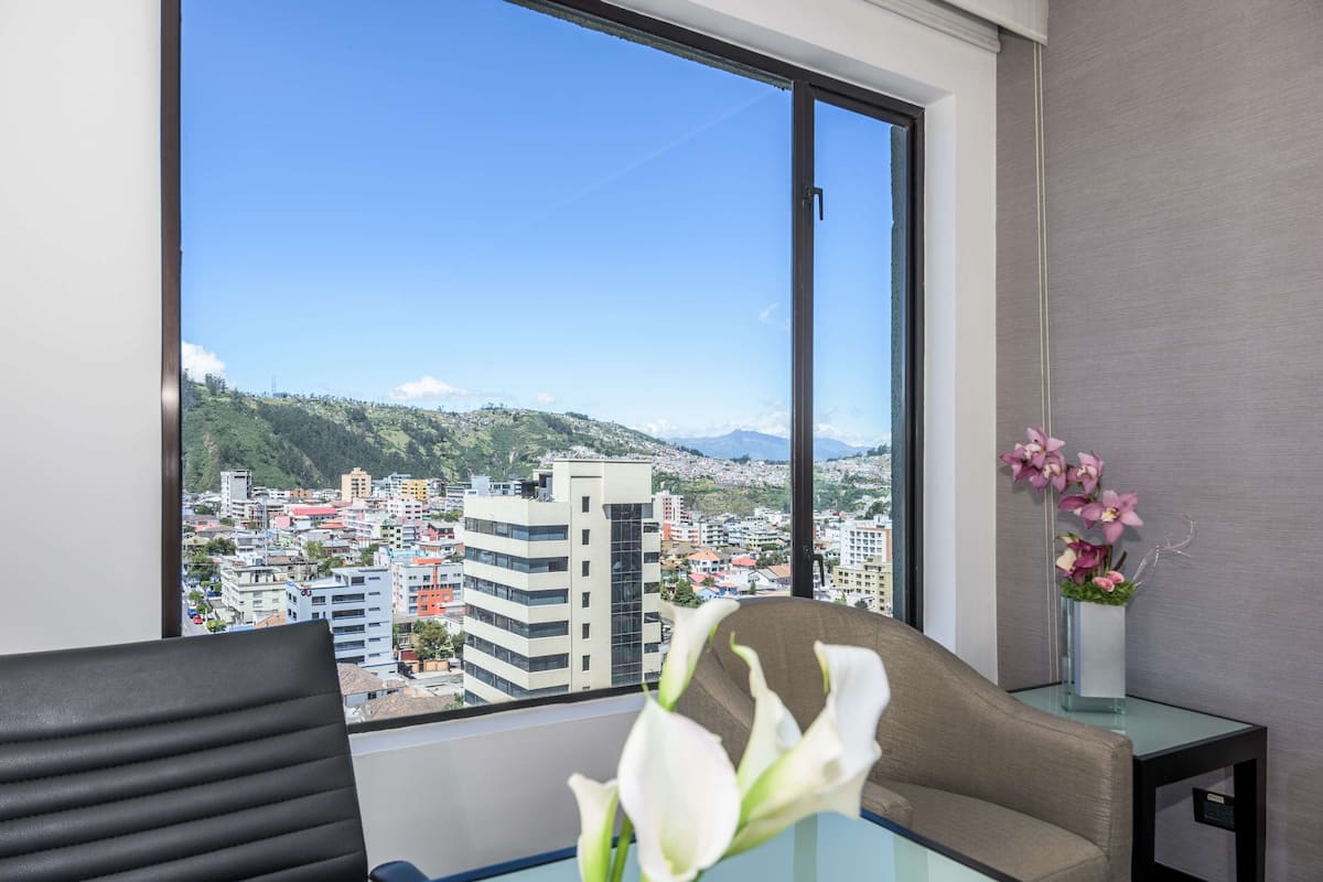 A hotel room with a large window offering a city view, a glass table, a chair, and a vase with flowers.