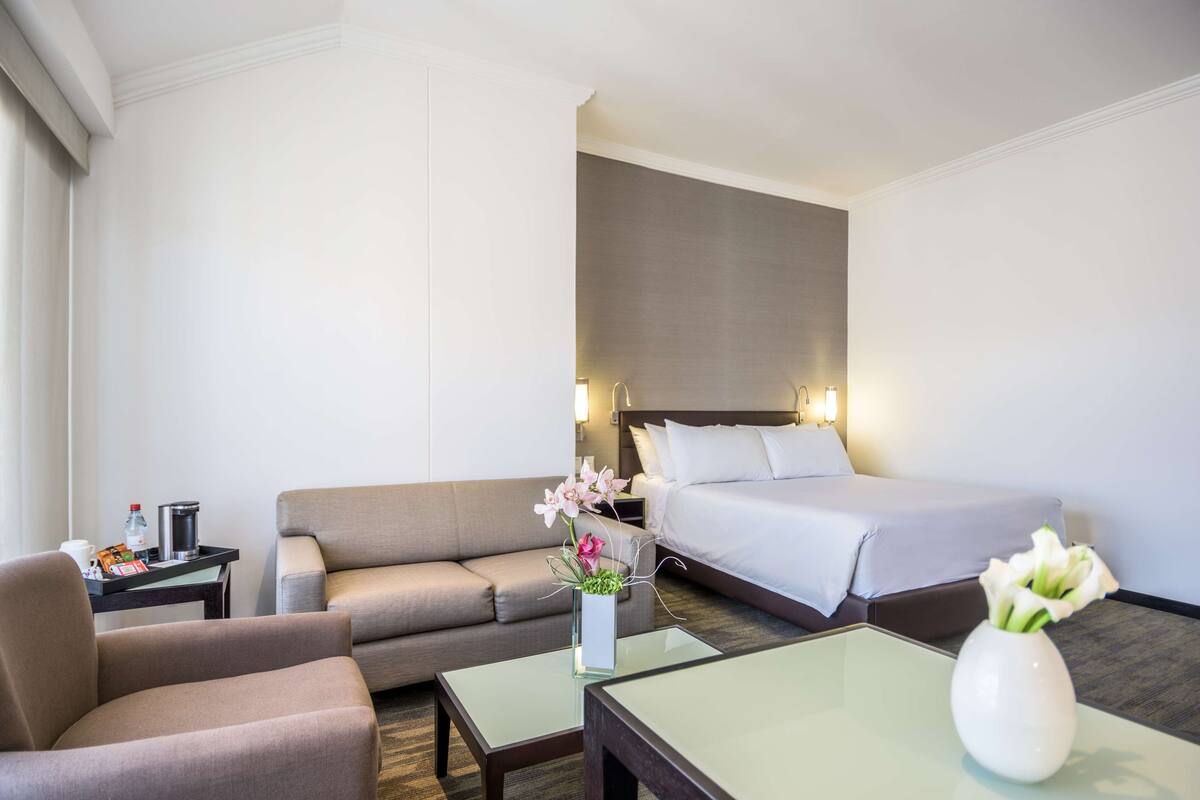A hotel room with a bed, a sofa, a coffee table, and a small table with a vase of flowers.