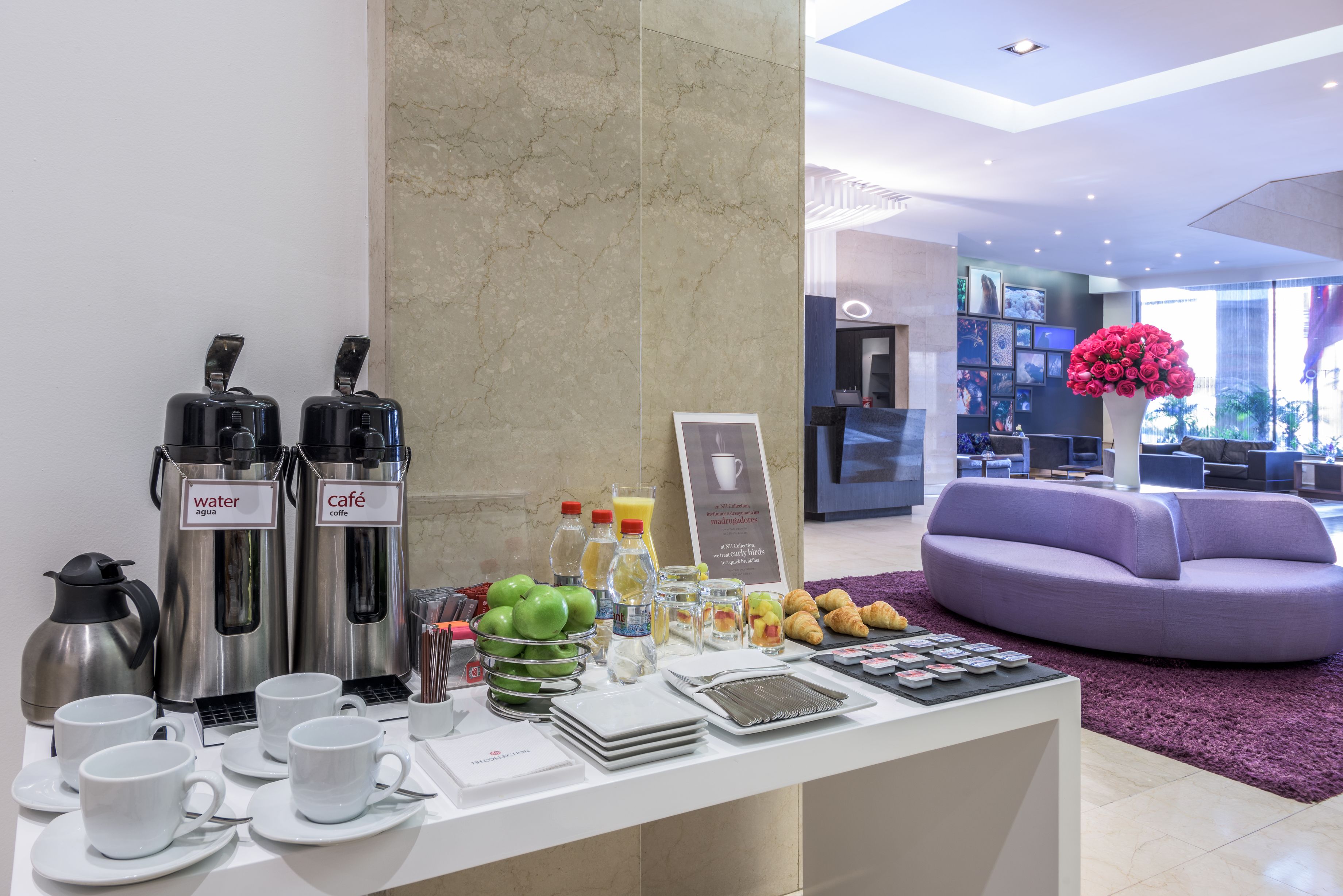 Una mesa de desayuno con dispensadores de café y jugo, un frutero y una exposición de cruasanes.