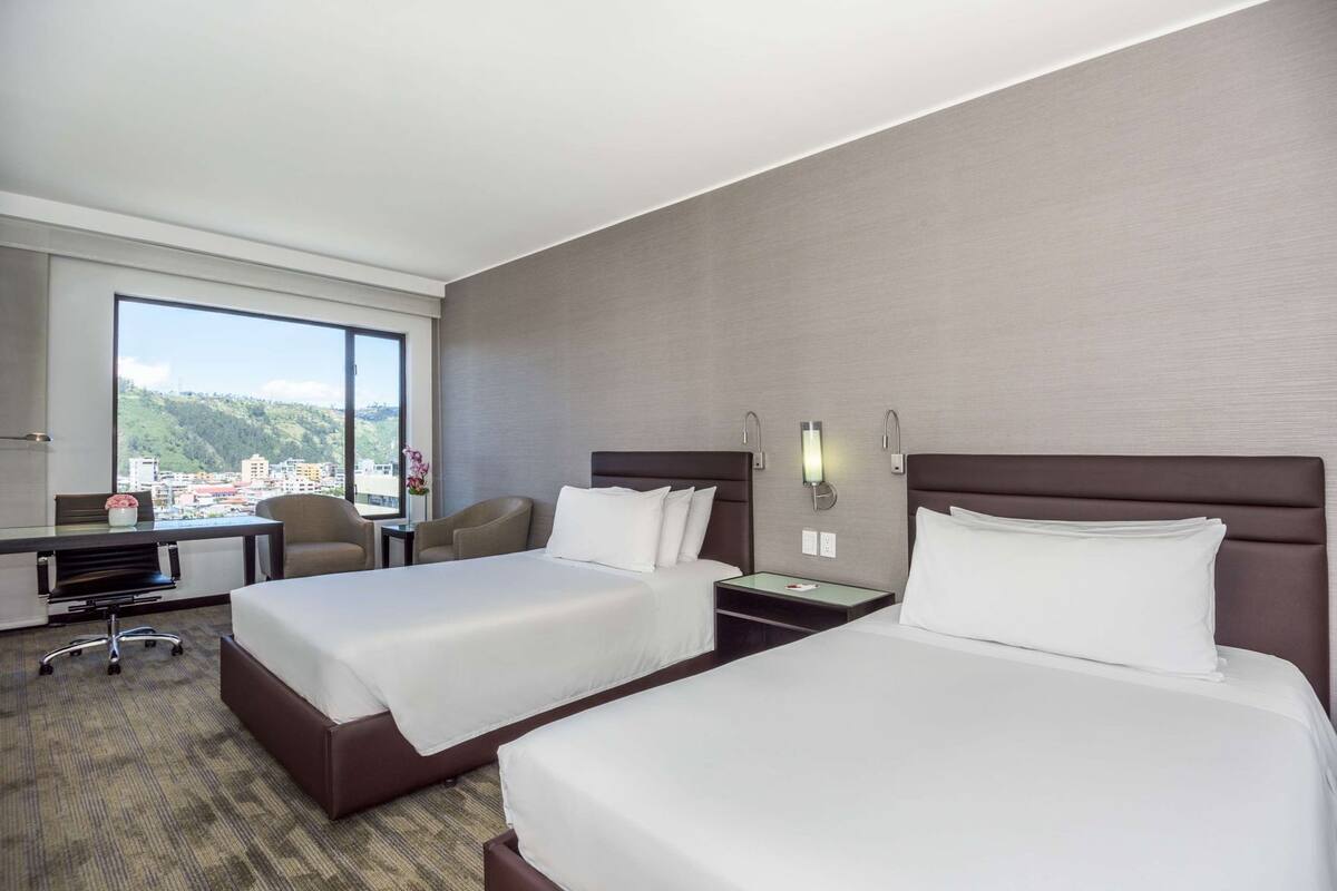 A hotel room with two beds, a desk, a chair, and a large window offering a city view.