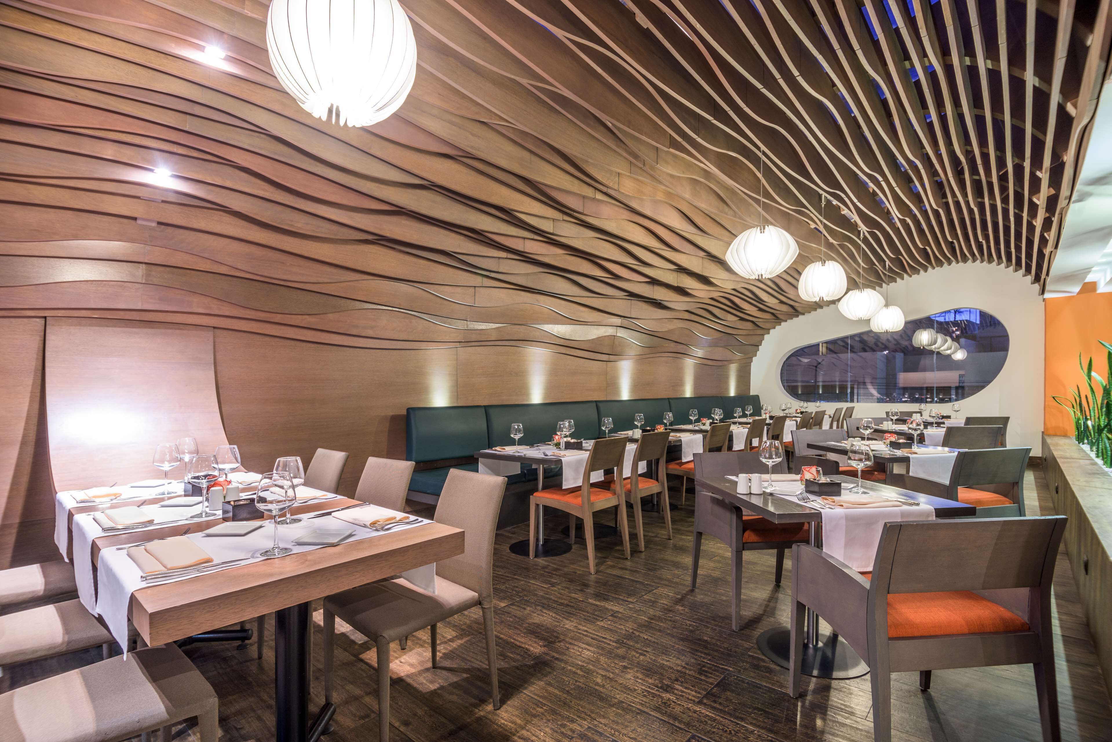 A modern restaurant with wooden ceiling panels, round pendant lights, and a green booth area. Tables are set with wine glasses and menus.