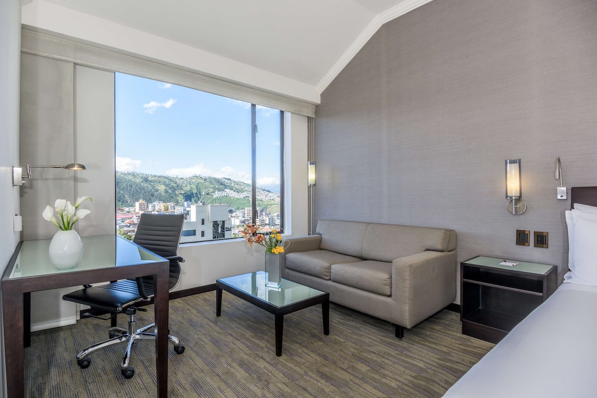 A hotel room with a desk, office chair, sofa, and a nightstand with a lamp.