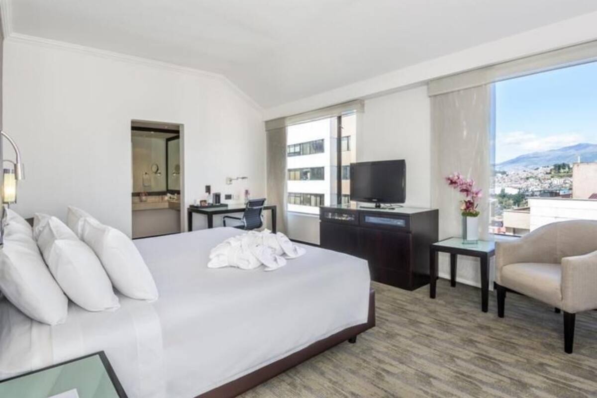 A hotel room with a bed, a desk, a television, a chair, and a view of the city.