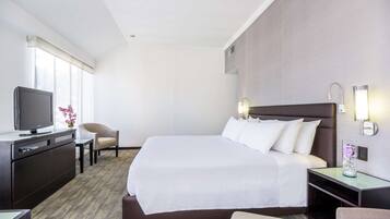 Habitación de hotel con una cama grande, un escritorio con televisor, una silla y una mesita de noche con lámpara.