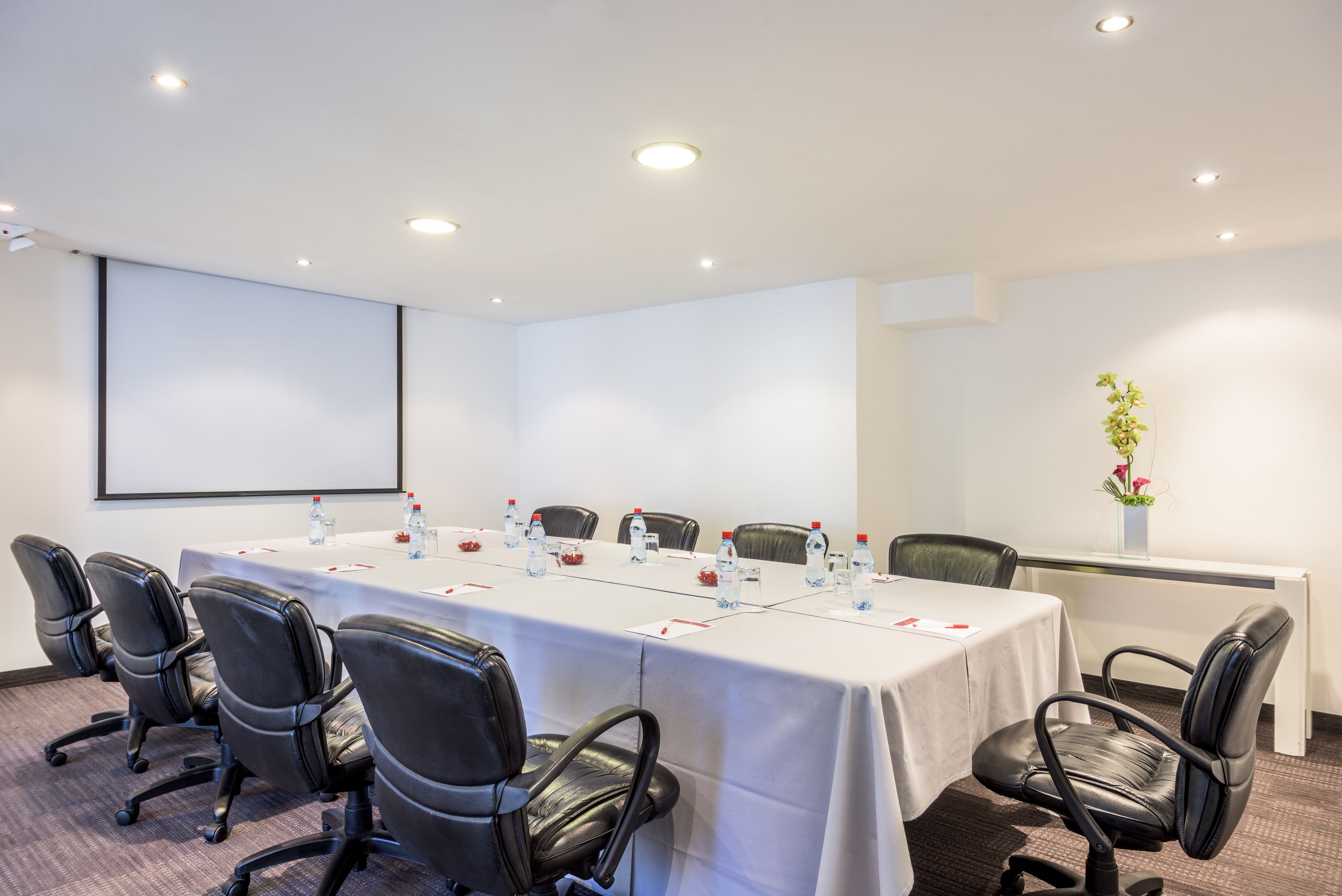 Un salón de conferencias con una larga mesa, sillas de cuero negro, botellas de agua y una pantalla de proyector.