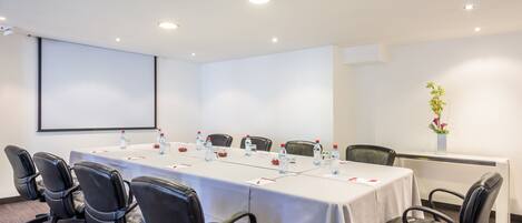 Un salón de conferencias con una larga mesa, sillas de cuero negro, botellas de agua y una pantalla de proyector.