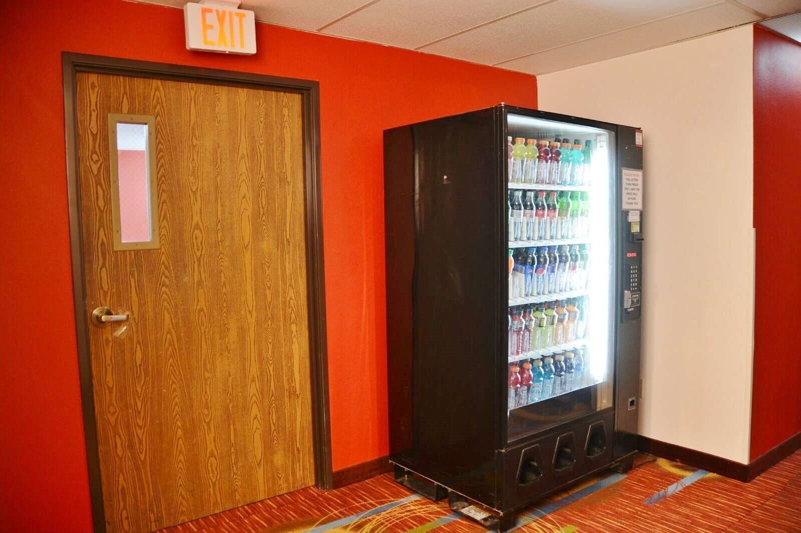 A hotel room with a wooden door, an exit sign, and a vending machine with various beverages.