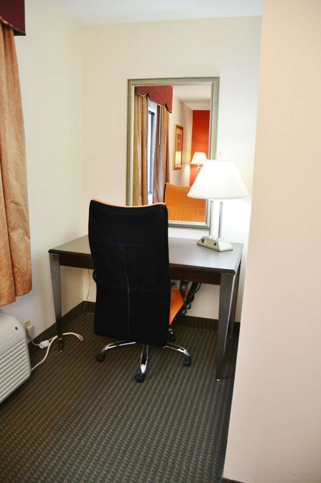 A hotel room with a desk, a chair, a lamp, and a mirror.
