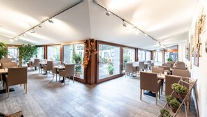 A spacious indoor area with multiple tables and chairs, large windows, and indoor plants. - Hotel Sauerlacher Post (Sauerlach)