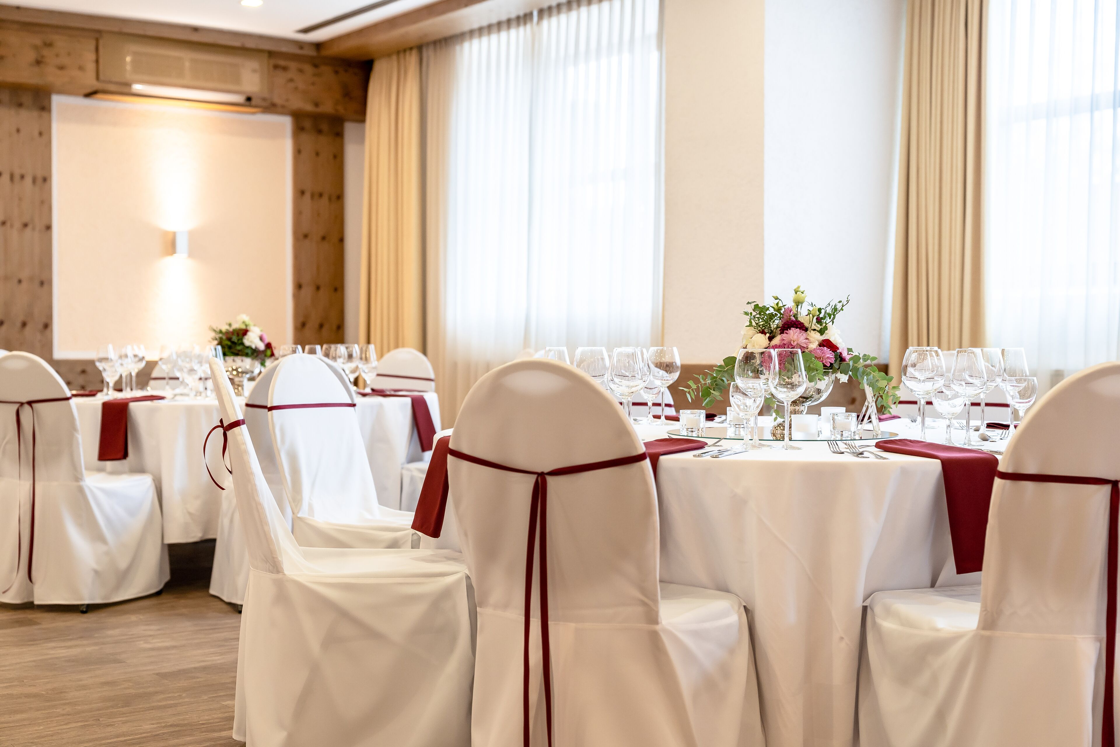 A banquet hall with round tables set for a formal event, white chairs with red sashes, and a large window with curtains.