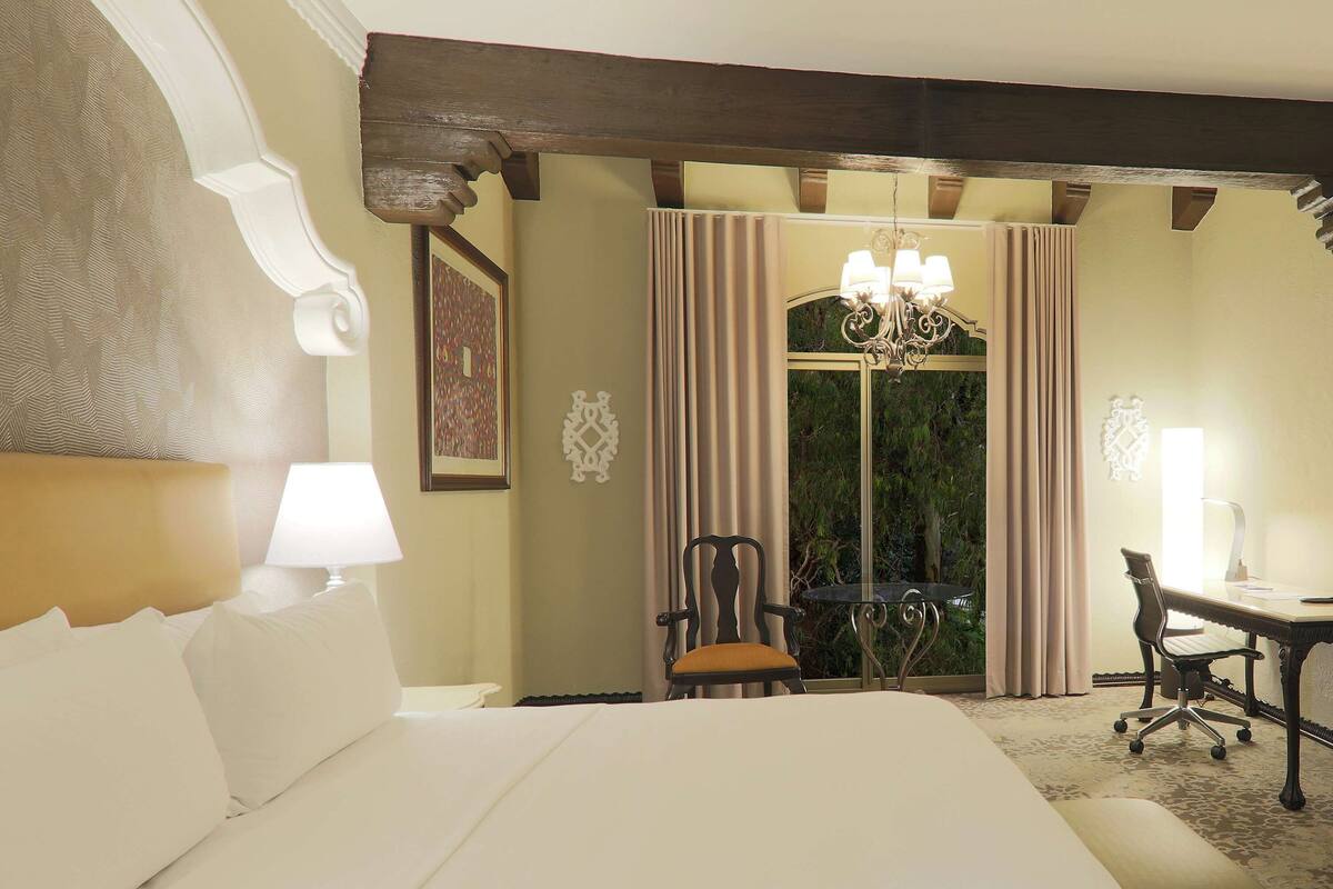 Een hotelkamer met een groot bed, een bureau met stoel, een kroonluchter en uitzicht op het omliggende landschap.