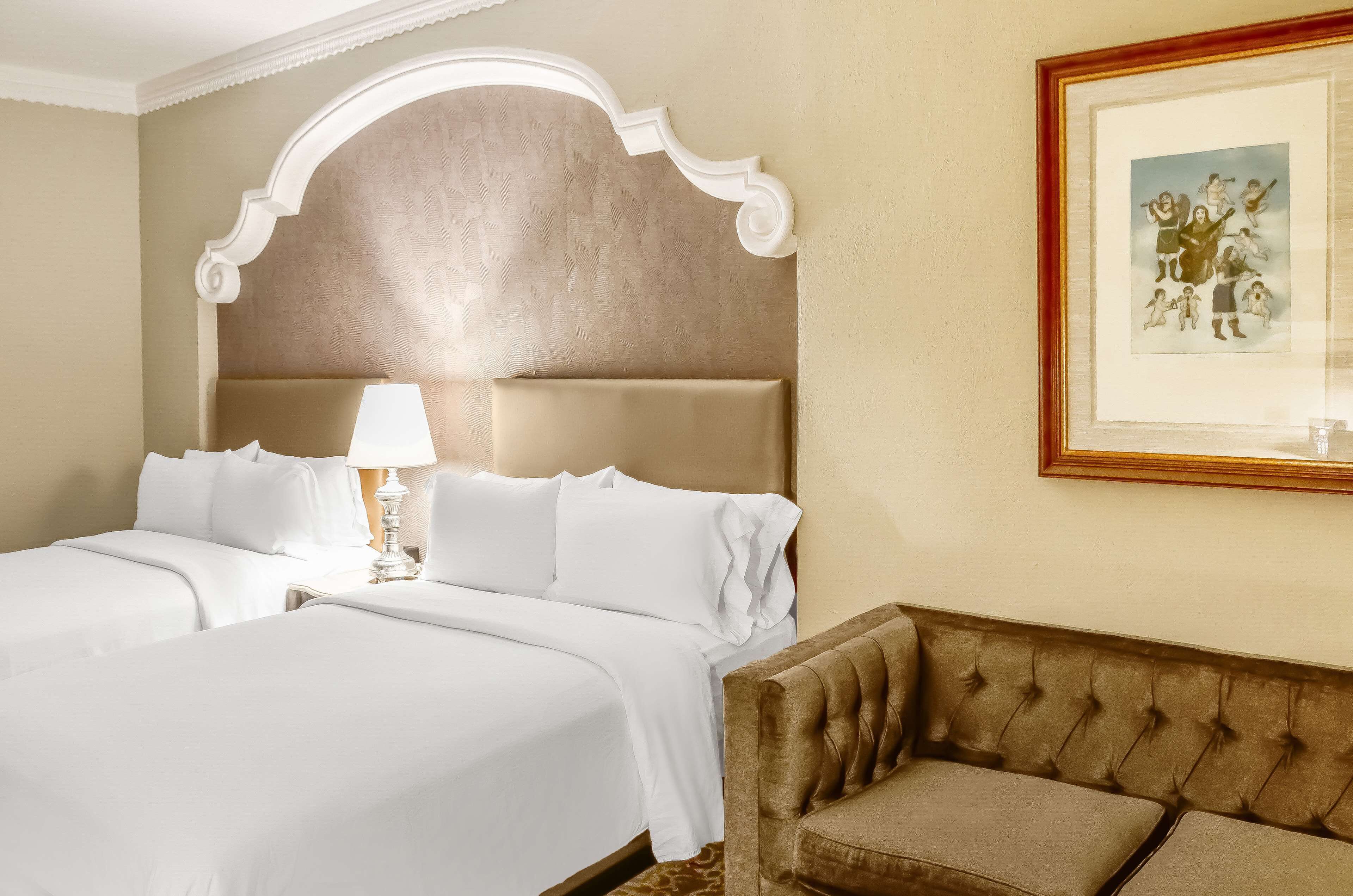 A hotel room with two beds, a large headboard, a framed picture on the wall, and a brown sofa.