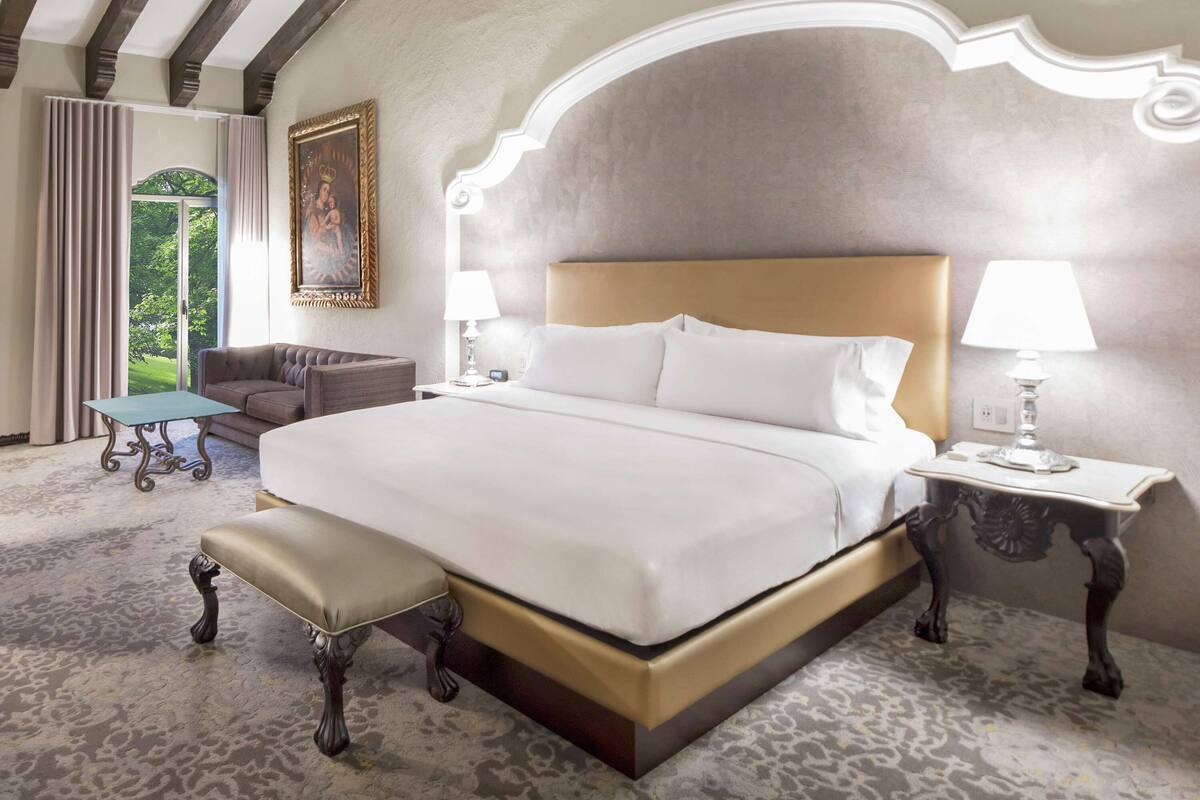 Een hotelkamer met een groot bed, een bankje, een nachtkastje met lamp, een stoel en een schilderij aan de muur.