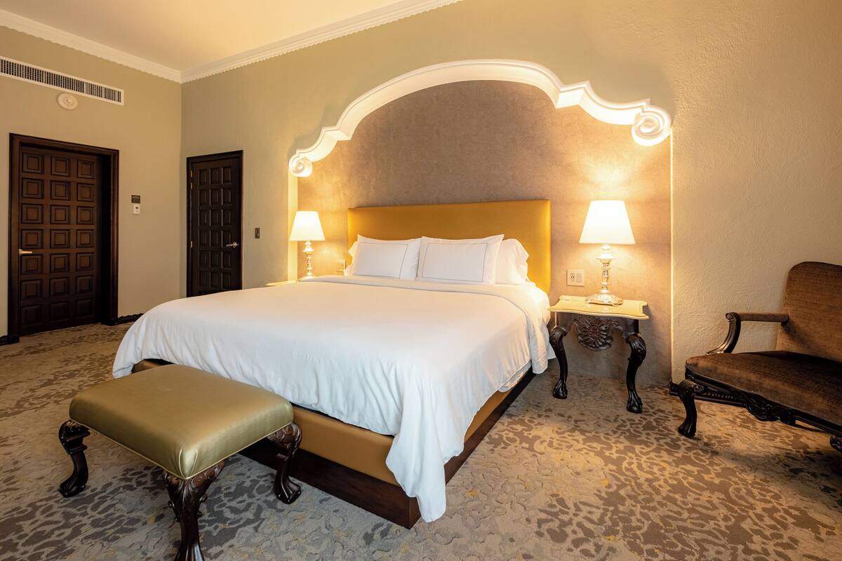 Een hotelkamer met een groot bed, twee bedlampen, een houten bankje en een stoel.