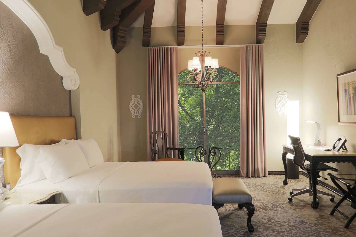 Een hotelkamer met een groot bed, een bureau en een stoel. Er hangt een kroonluchter aan het plafond en er is een raam met gordijnen.