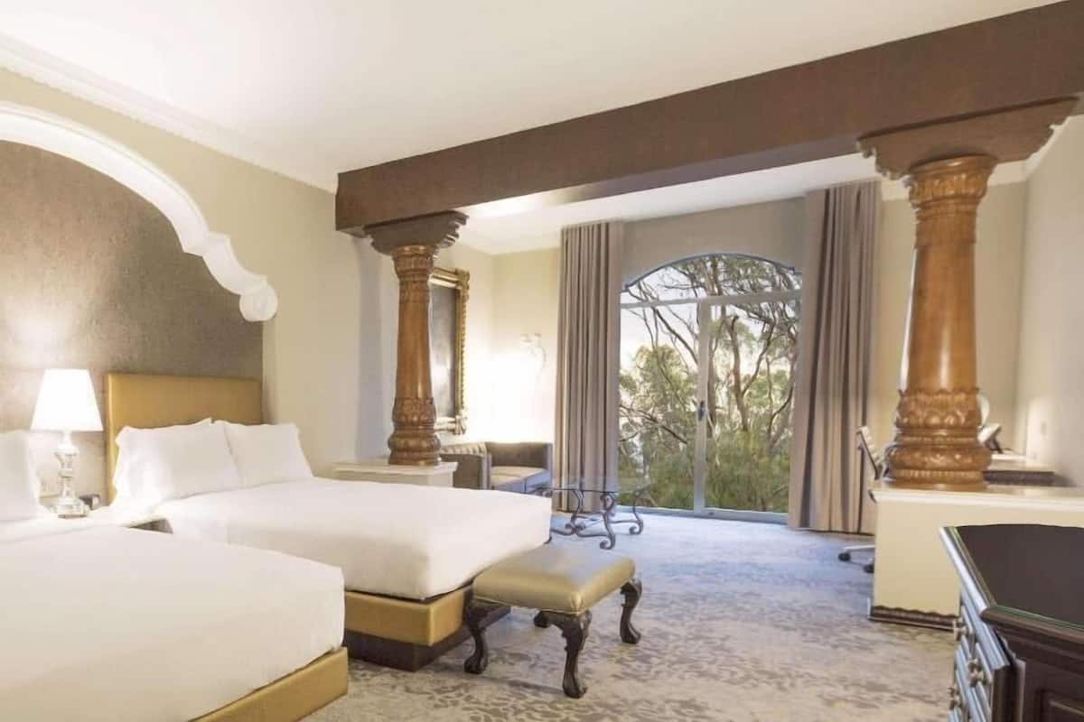 Een hotelkamer met twee bedden, een bureau, een stoel en een groot raam met uitzicht op bomen.