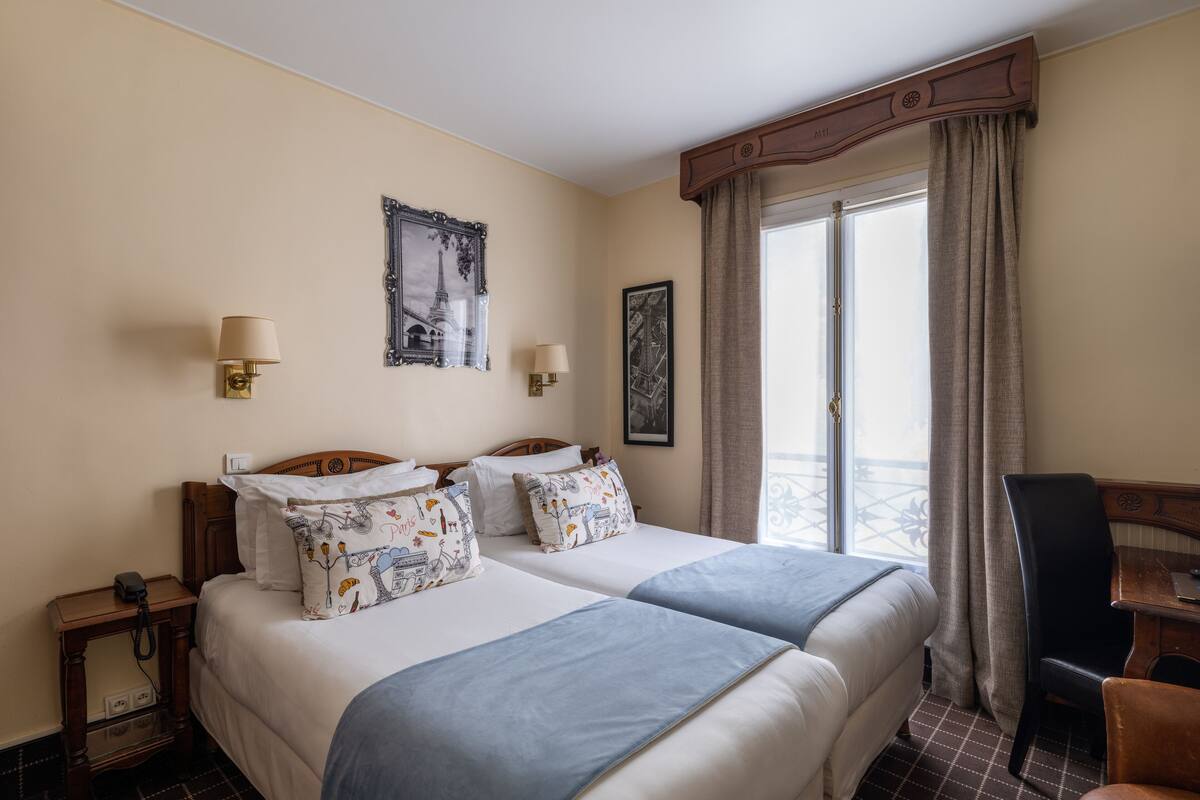 A hotel room with two beds, a wooden headboard, a desk, a chair, a telephone, and a window with curtains.