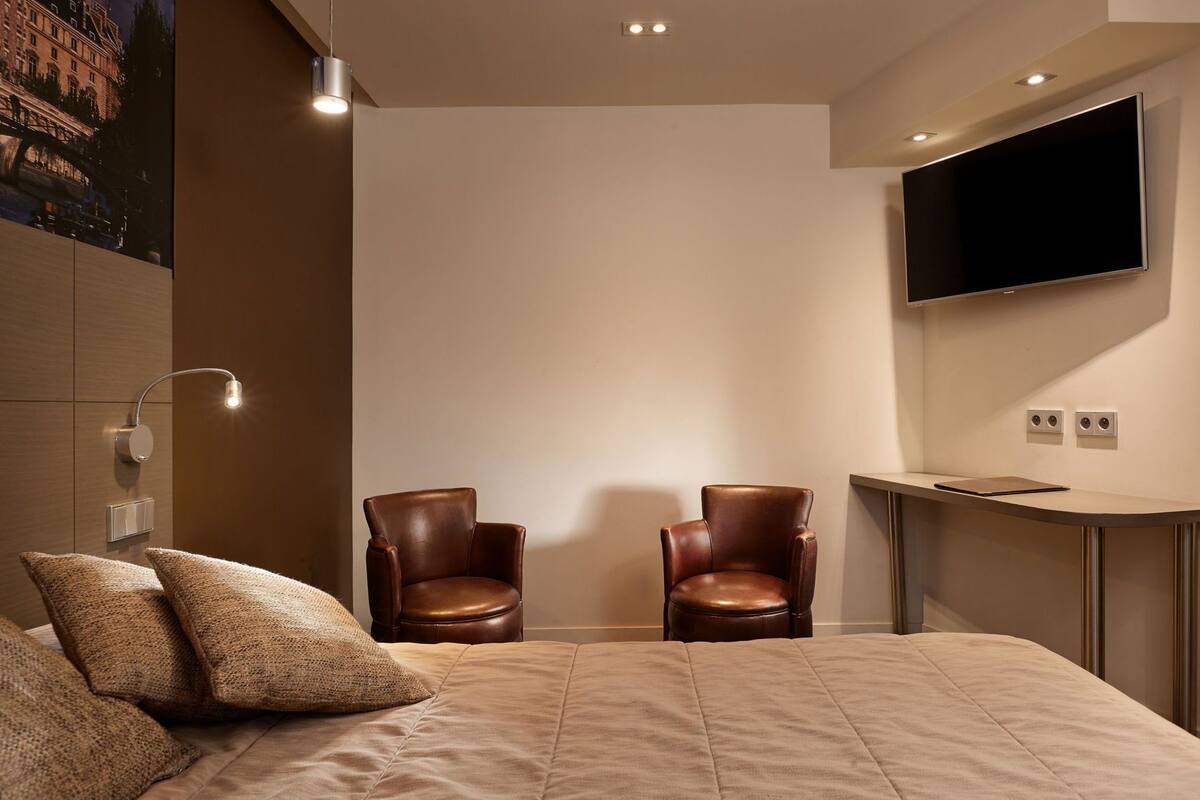 A modern hotel room with a large bed, two leather armchairs, a flat-screen TV, and a desk with a tablet.