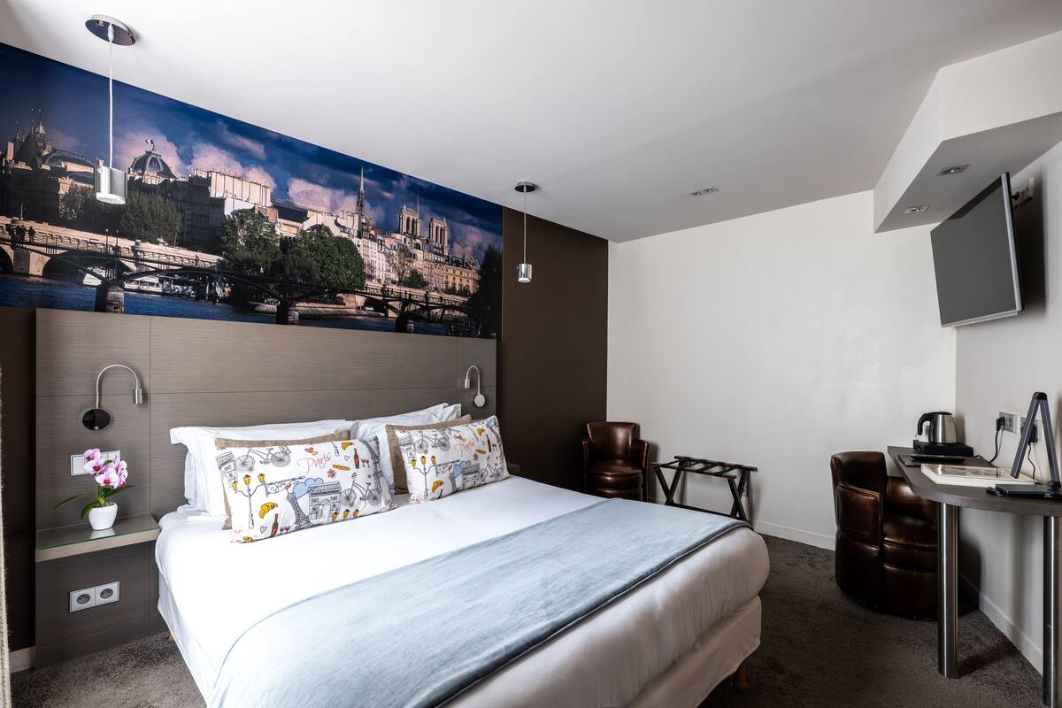 A hotel room with a large bed, a desk with a chair, a TV, and a mural of a cityscape on the wall.