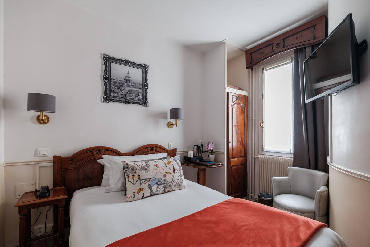 A hotel room with a bed, bedside tables, a desk, a chair, a TV, and a window with curtains.