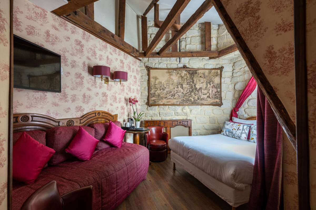 A bedroom with a wooden ceiling, a stone wall, a bed, a sofa, and a table with a lamp.