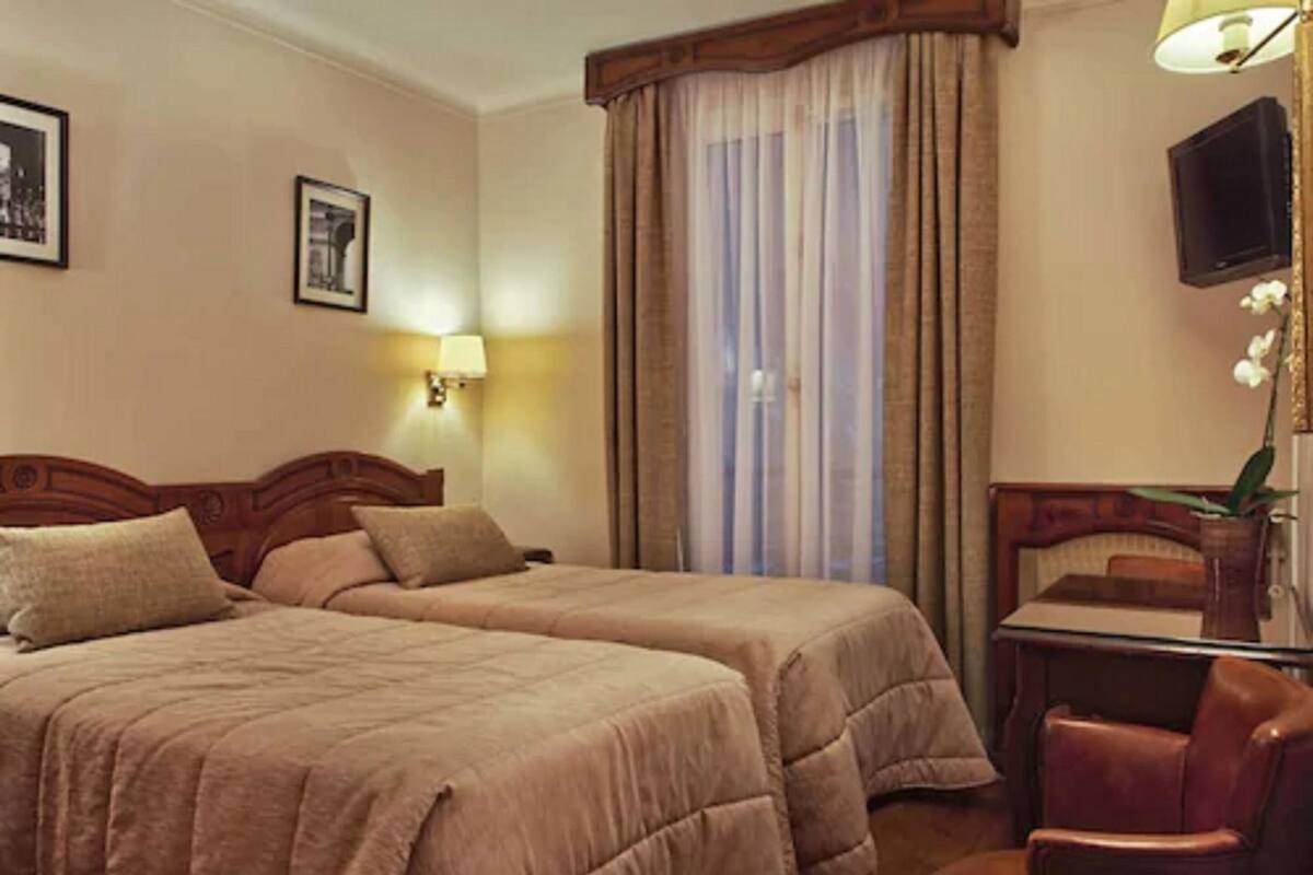 A hotel room with two beds, a desk, a chair, a TV, and a window with curtains.